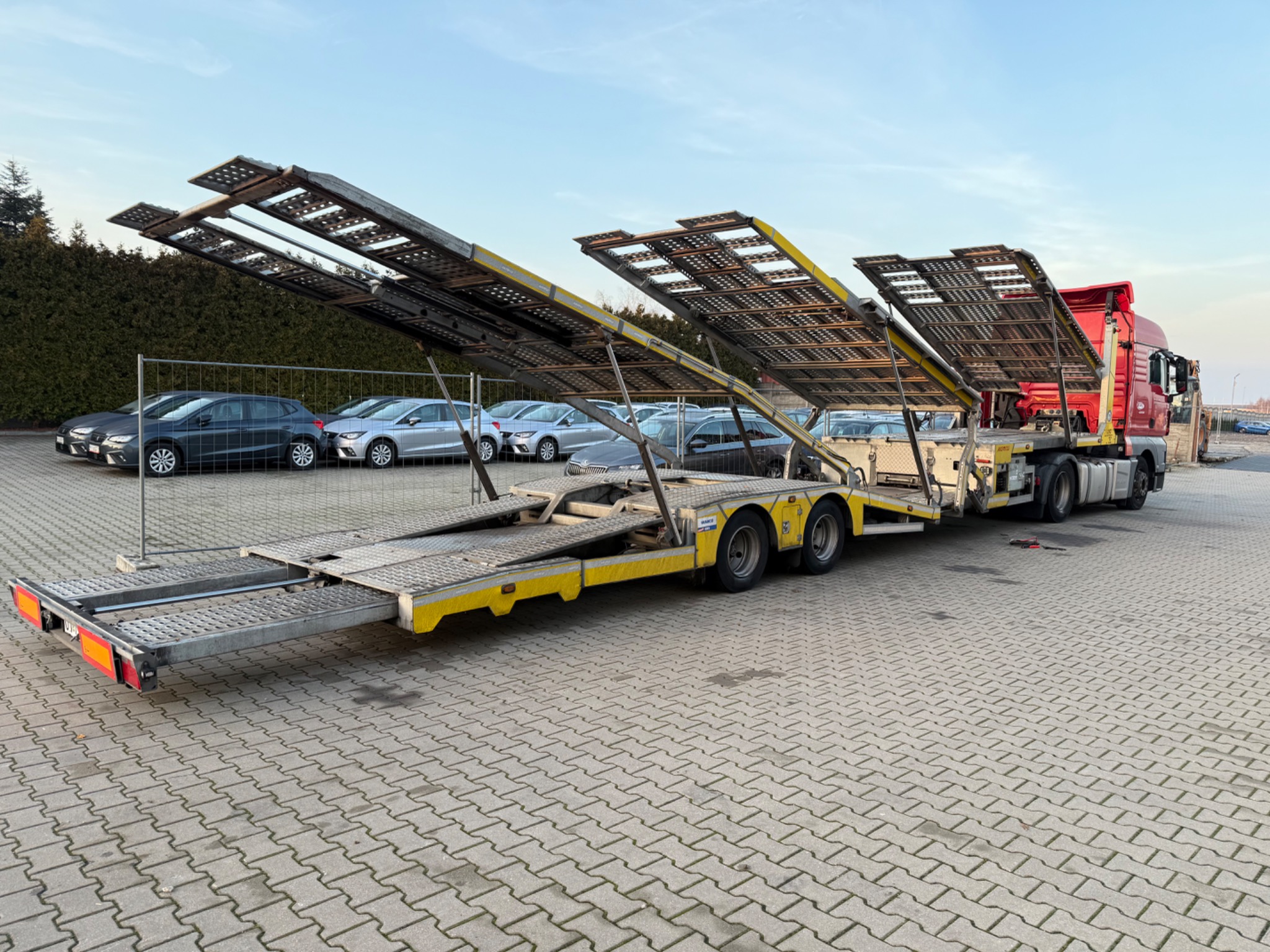 Czerwona ciężarówka z żółtą lawetą na parkingu, z podniesionymi platformami załadunkowymi. W tle zaparkowane samochody osobowe i zielony żywopłot.