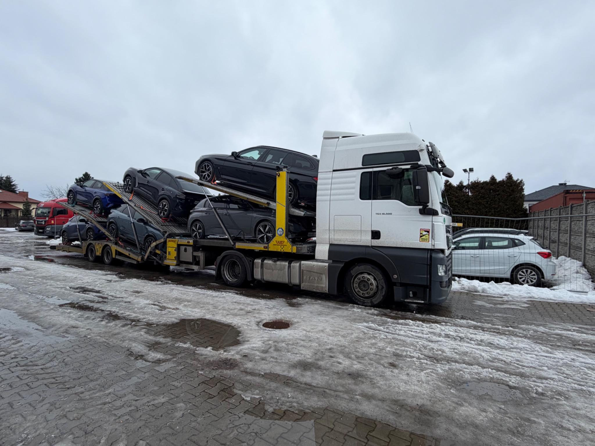 Laweta MAN z załadowanymi samochodami osobowymi, zaparkowana na placu w zimowej scenerii. Widoczny śnieg i błoto pośniegowe.