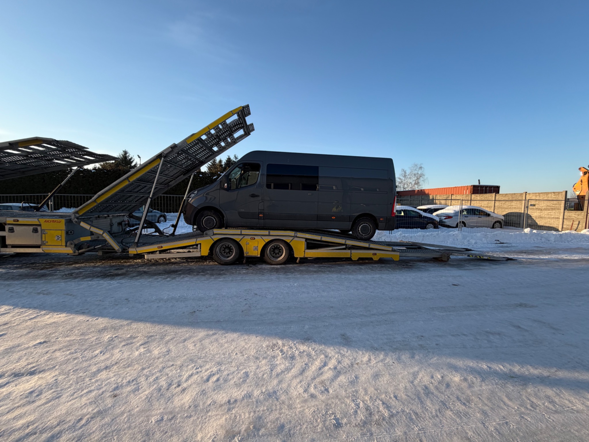 Szary bus na żółtej lawecie w Dawidach, gotowy do transportu. Widoczna rampa załadunkowa i zimowy krajobraz. Błękitne niebo w tle.
