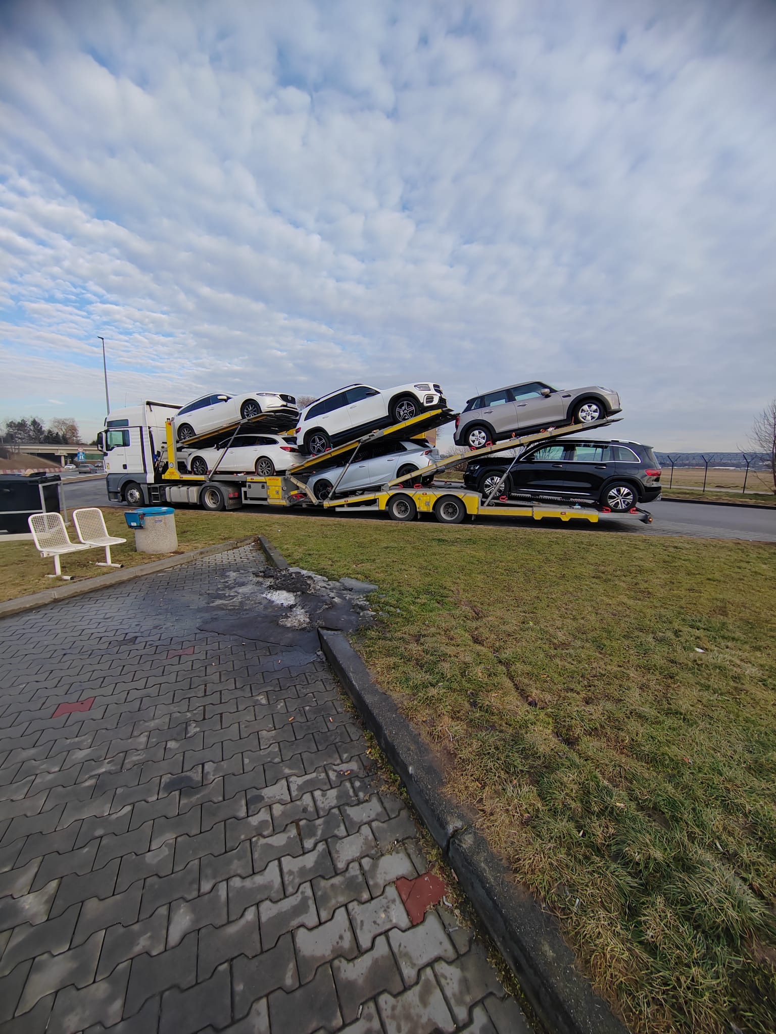 Autotransporter z kilkoma samochodami osobowymi na parkingu, widok z boku. Biała kabina ciągnika i żółta laweta. Pochmurne niebo w tle.