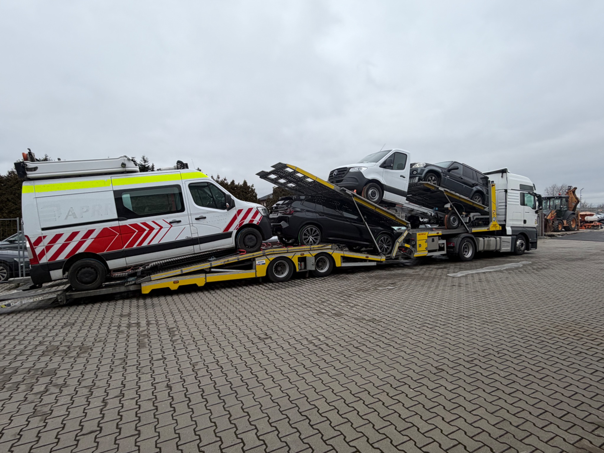 Wielopoziomowa laweta z trzema pojazdami, w tym ambulansem, na parkingu w Dawidach. Transport aut z zagranicy na specjalistycznej platformie. Szary dzień.