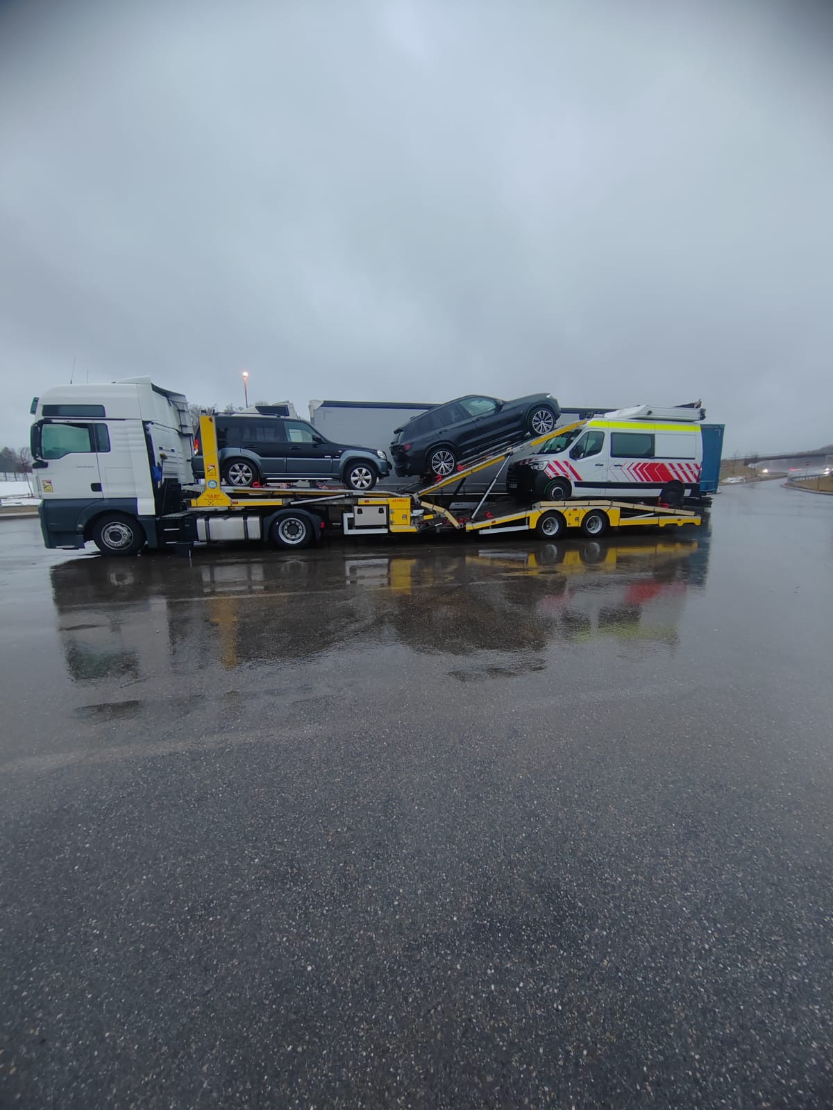 Ciężarówka laweta z trzema pojazdami: SUV, osobowy i ambulans. Transport aut na mokrym asfalcie, pochmurne niebo w tle. Widok z boku, lekki deszcz.