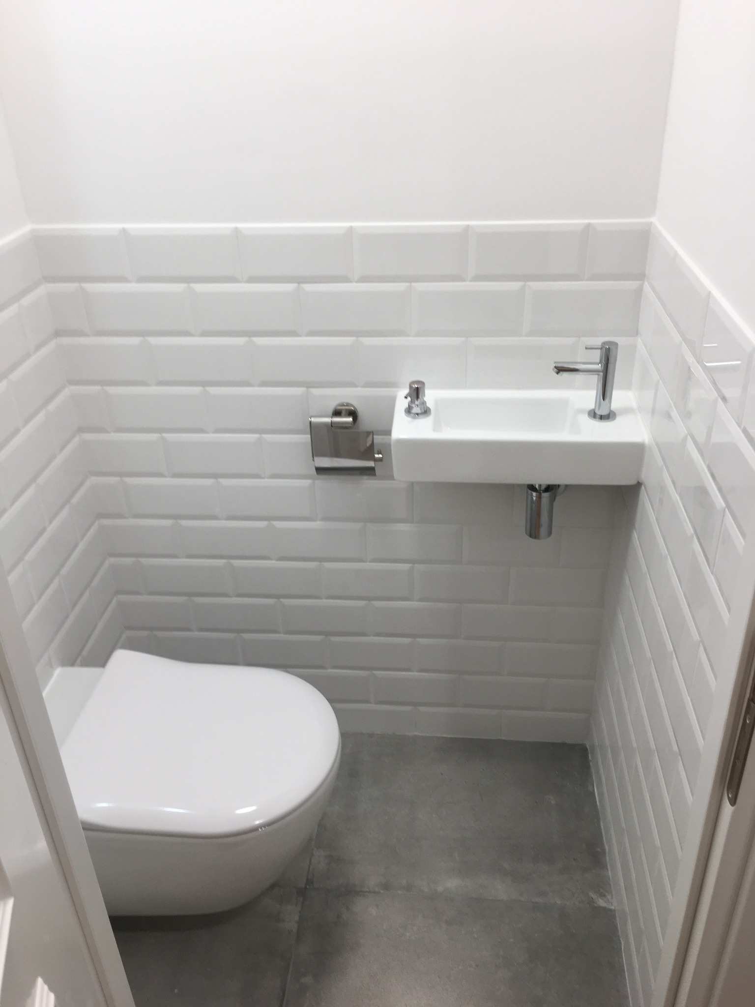 Mała, minimalistyczna łazienka w bieli: toaleta podwieszana, umywalka narożna z chromowaną armaturą, białe płytki typu metro i szara podłoga. Nowoczesne wykończenie.