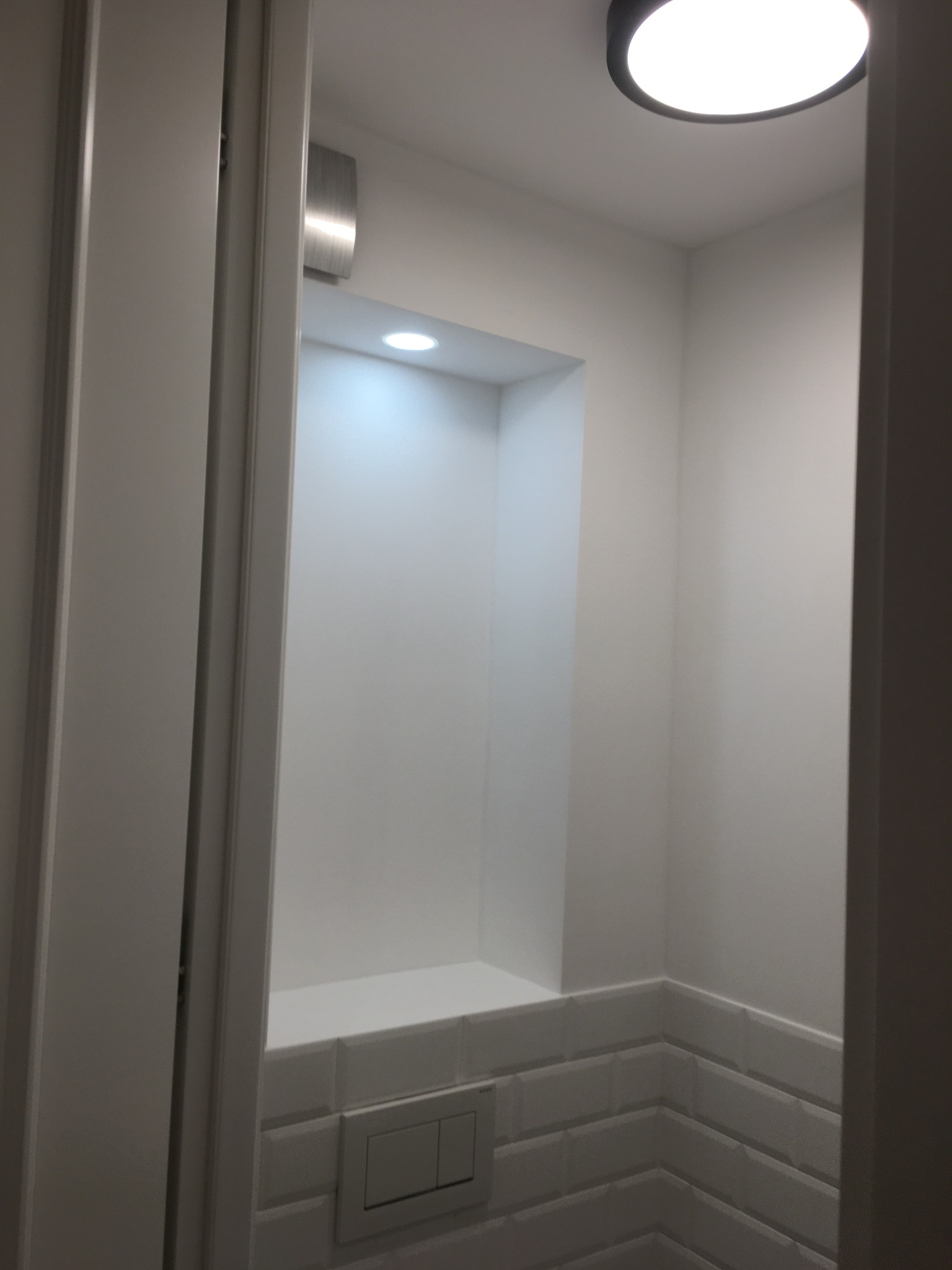 Nowoczesna, minimalistyczna łazienka z wnęką oświetloną punktowo, wykończona białymi płytkami typu metro i podwieszaną miską WC. Widok zza uchylonych drzwi.