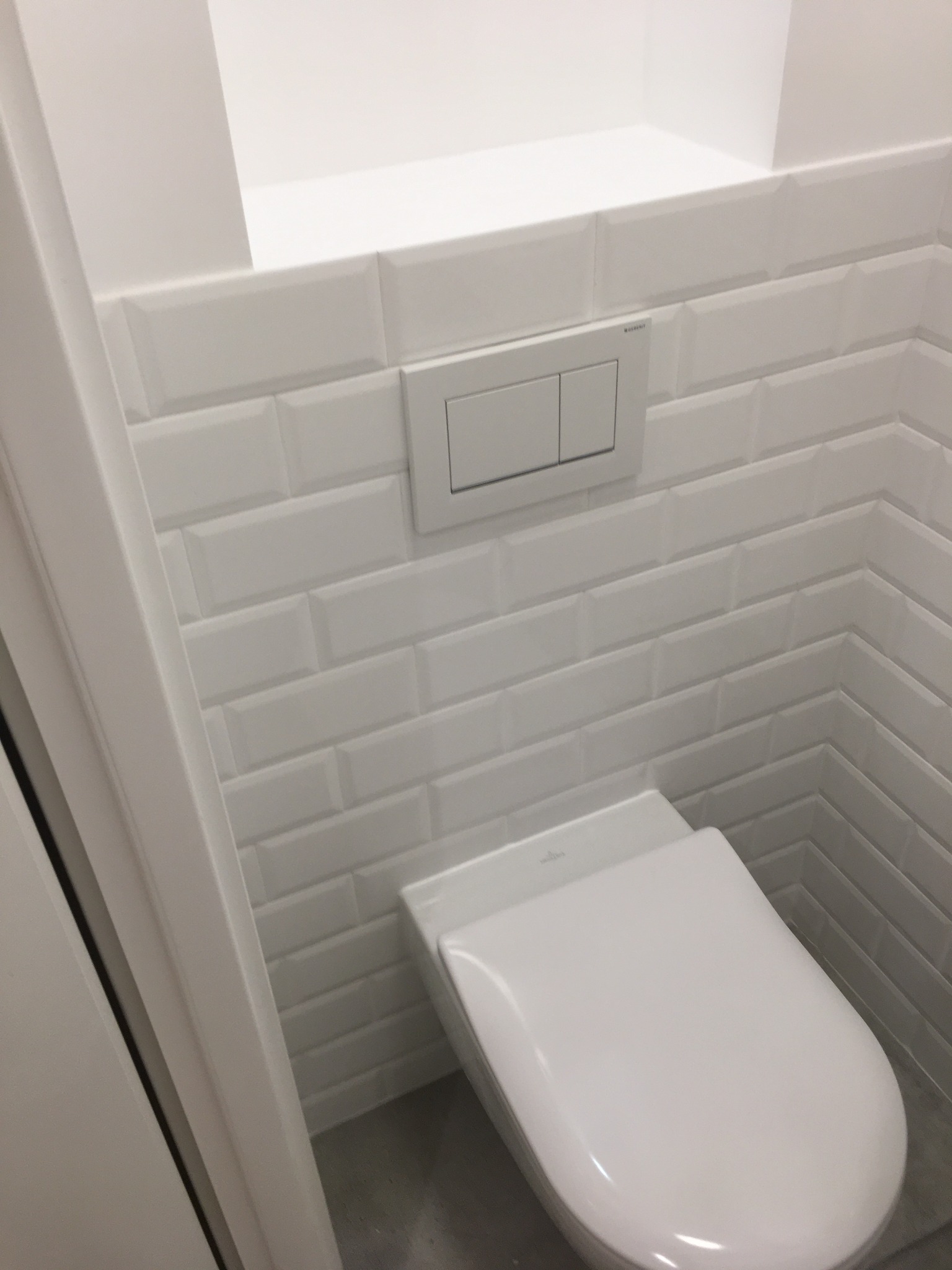 Biała toaleta podtynkowa z przyciskiem spłukującym na ścianie wyłożonej białymi, błyszczącymi płytkami typu metro w łazience. Minimalistyczny design.