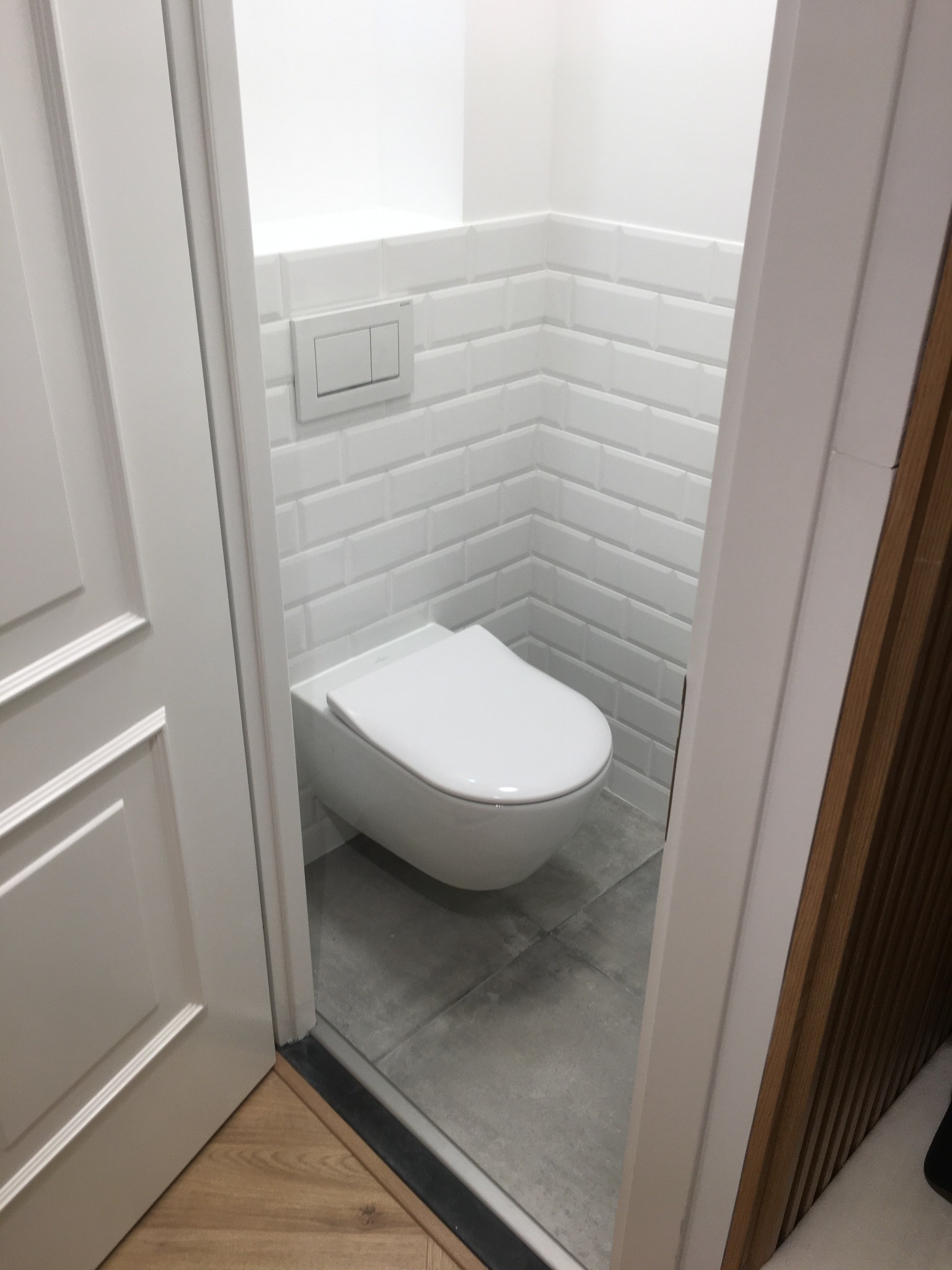 Nowoczesna toaleta podwieszana w wykończonej łazience. Białe płytki imitujące cegłę, szara podłoga, minimalistyczny design. Widok zza uchylonych drzwi.