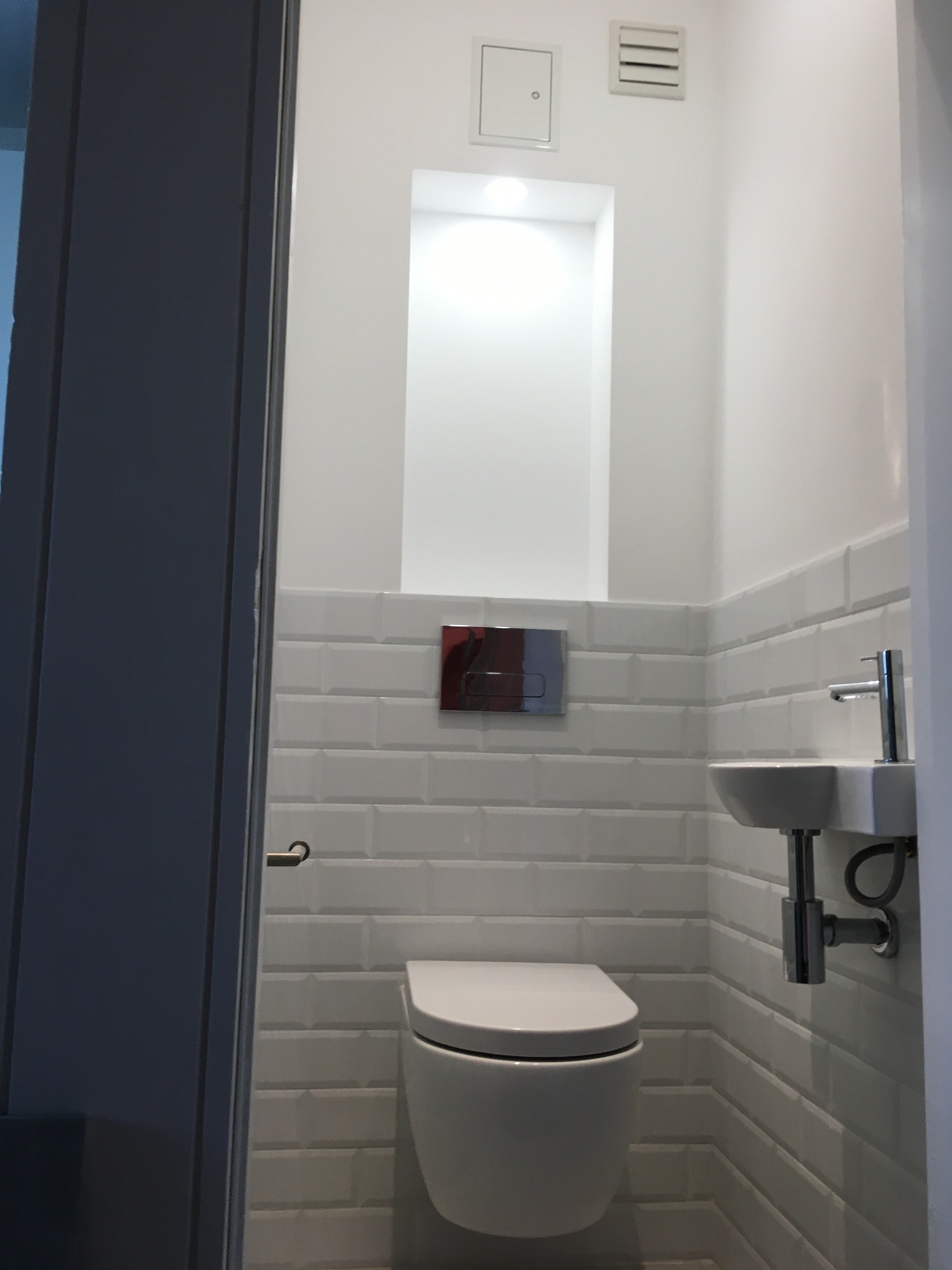 Nowoczesna, biała łazienka z toaletą podwieszaną, umywalką i białymi, prostokątnymi płytkami ułożonymi w cegiełkę. W ścianie wnęka z oświetleniem.