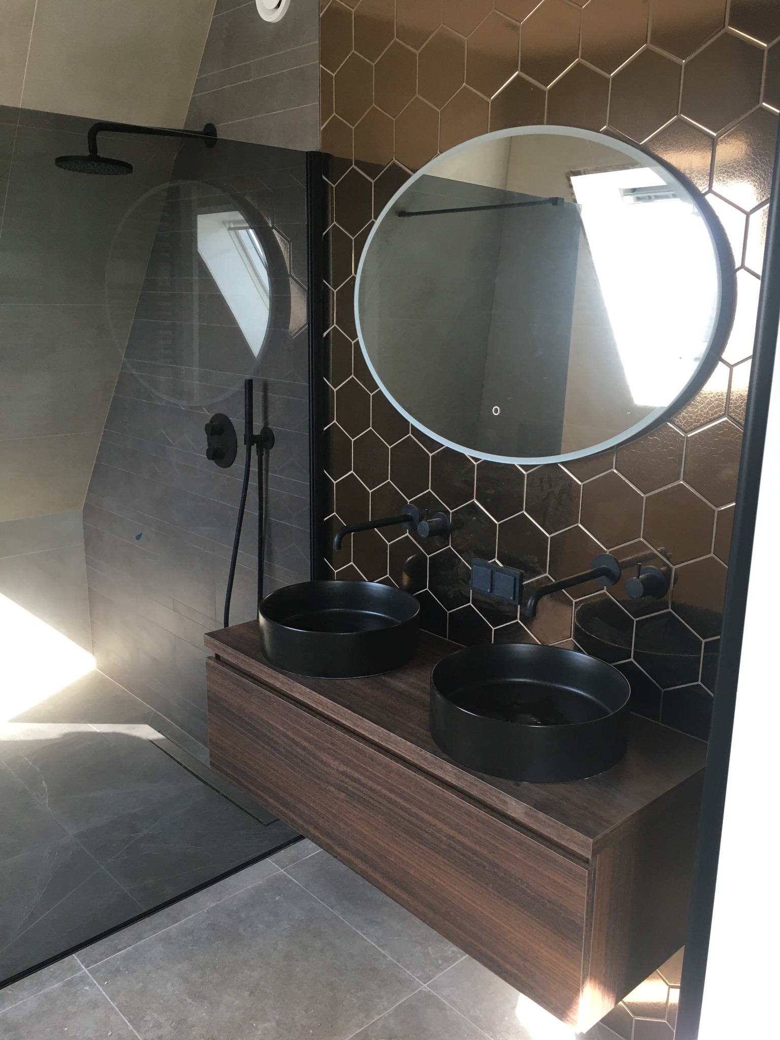 Nowoczesna łazienka z kabiną typu walk-in, umywalkami w czarnych misach na drewnianej szafce i lustrem w owalnej ramie na ścianie z heksagonalnych płytek.