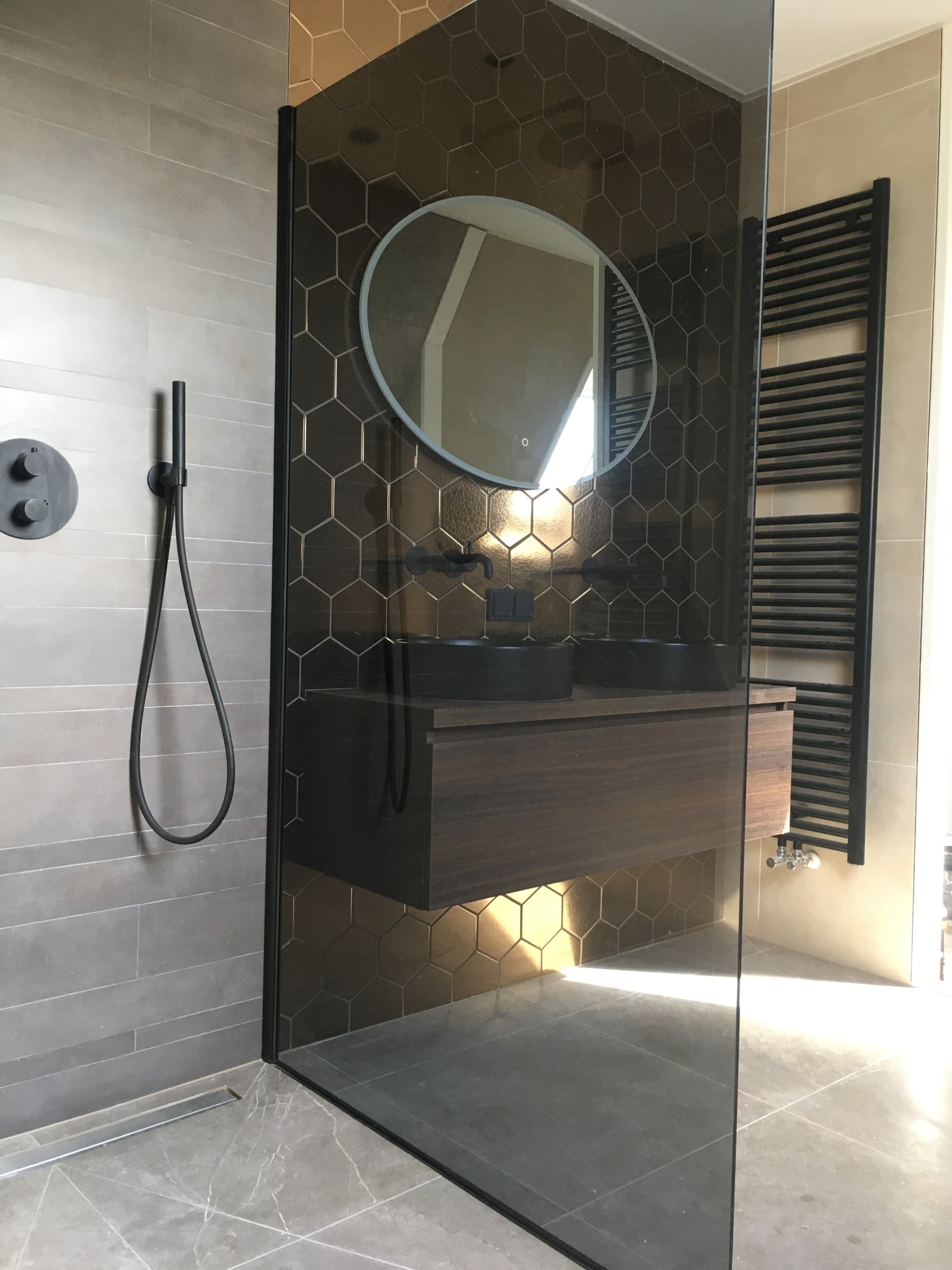 Nowoczesna łazienka z heksagonalnymi płytkami, szklaną kabiną prysznicową i podwieszaną szafką z umywalkami. Minimalistyczny design, czarne akcenty i subtelne oświetlenie.