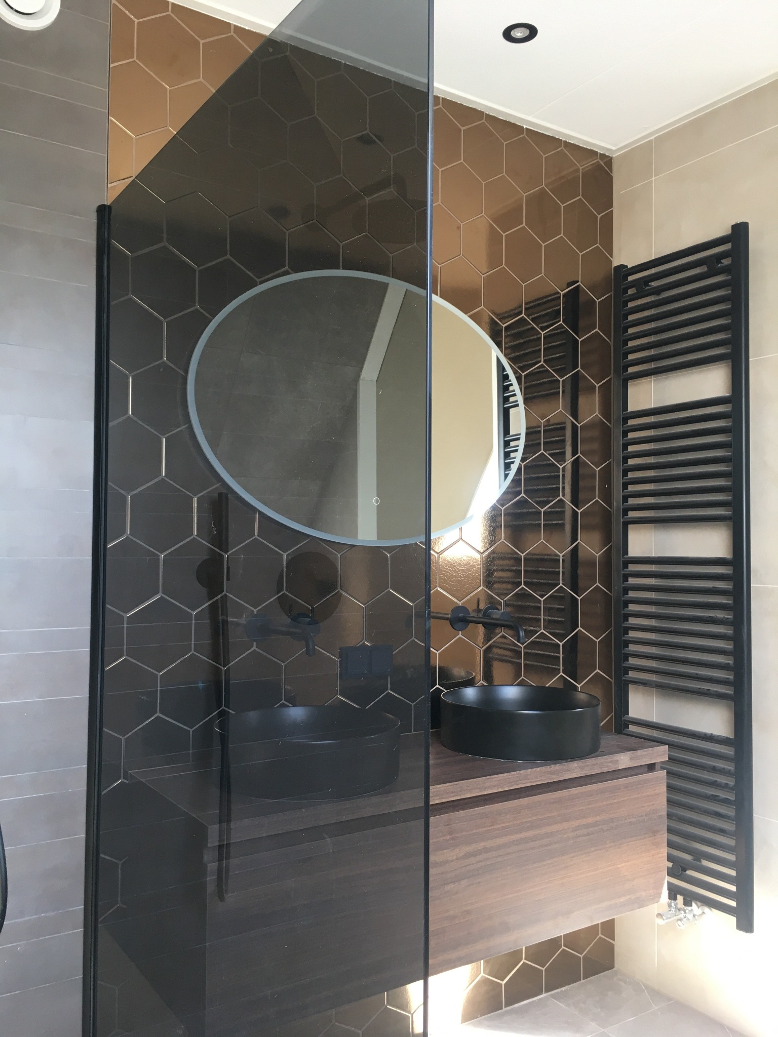 Nowoczesna łazienka z heksagonalnymi płytkami w odcieniach brązu, czarną armaturą i owalnym lustrem. Kabina prysznicowa ze szklaną ścianką.