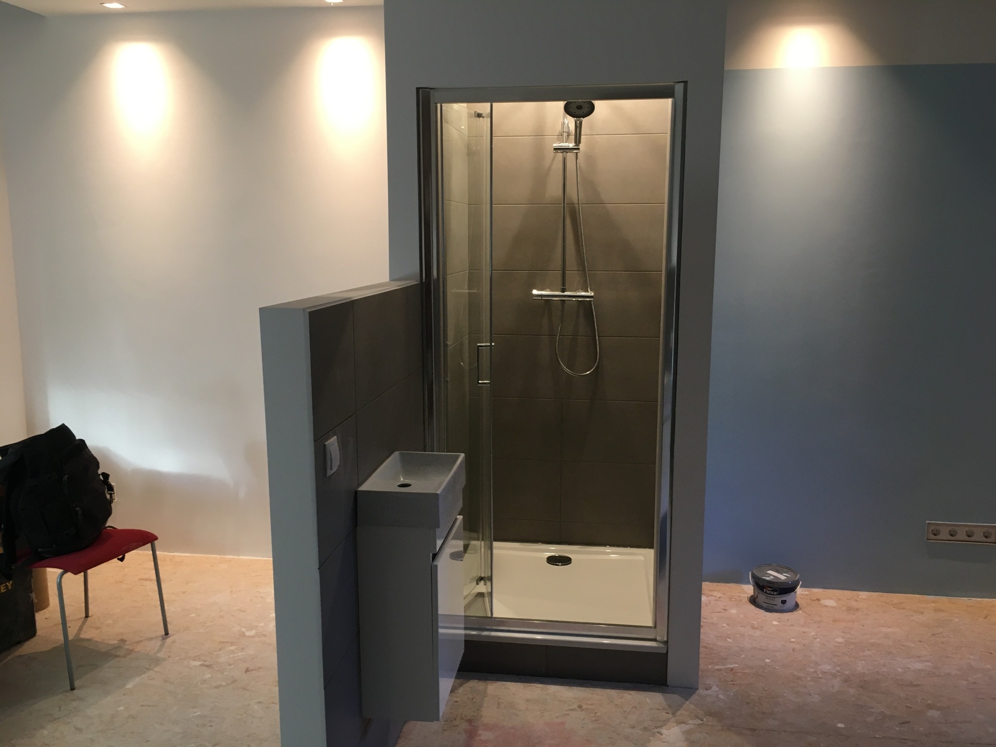 Nowoczesna kabina prysznicowa z umywalką w szarej łazience, widok z przodu. Na podłodze widoczne ślady po remoncie, obok puszka z farbą.