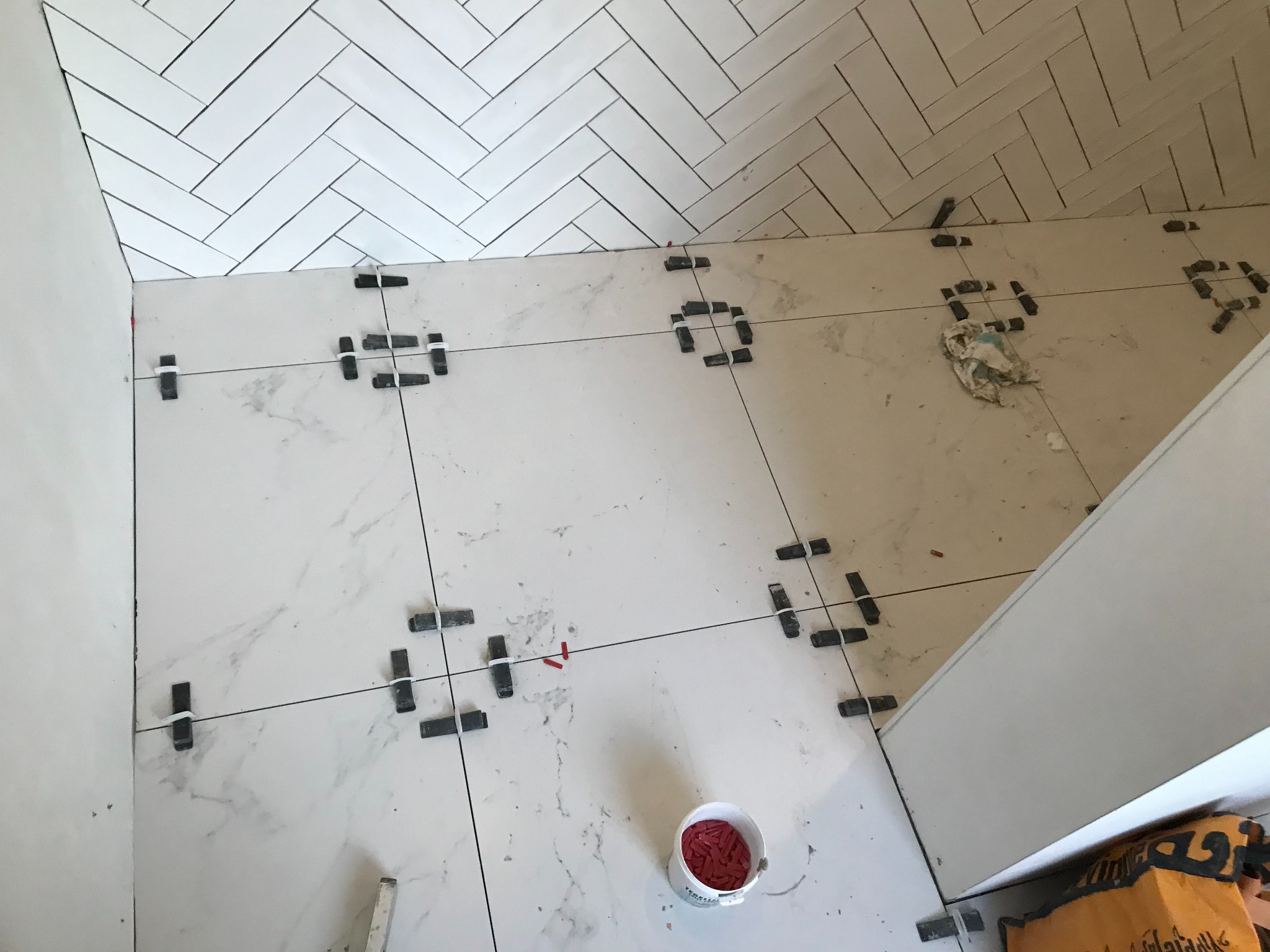 Układanie białych płytek imitujących marmur na podłodze i ścianie w jodełkę. Widoczne krzyżyki dystansowe i wiaderko z klinami. Etap prac wykończeniowych.