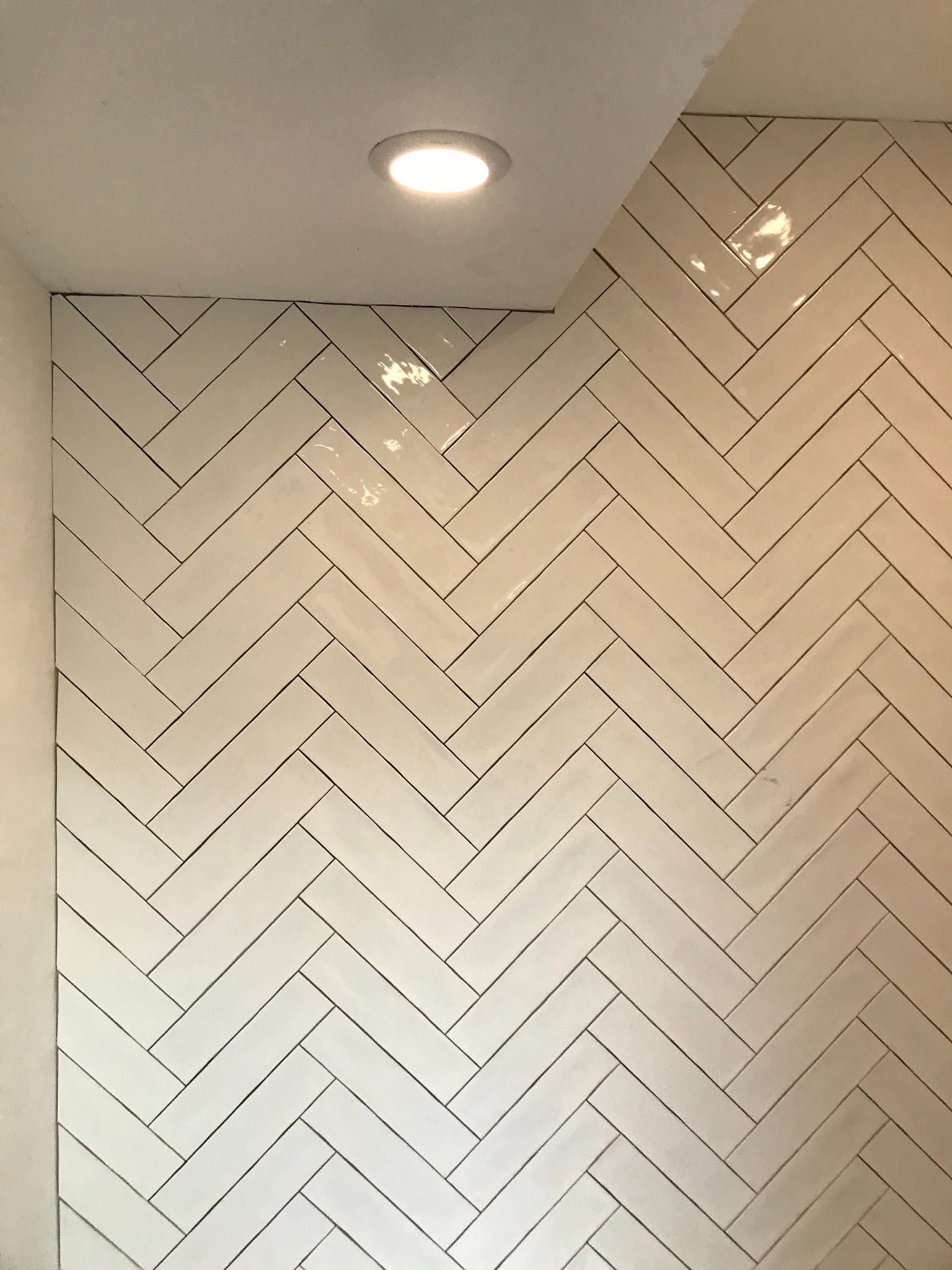 Ściana wyłożona białymi, błyszczącymi płytkami ułożonymi w jodełkę. Widoczna część sufitu z wbudowanym oświetleniem punktowym. Minimalistyczny design.