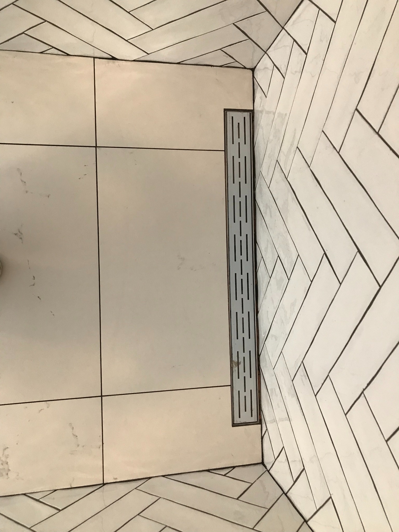 Białe płytki w jodełkę i kwadraty z odpływem liniowym w kabinie prysznicowej. Minimalistyczny design, jasne wnętrze, precyzyjne ułożenie płytek.