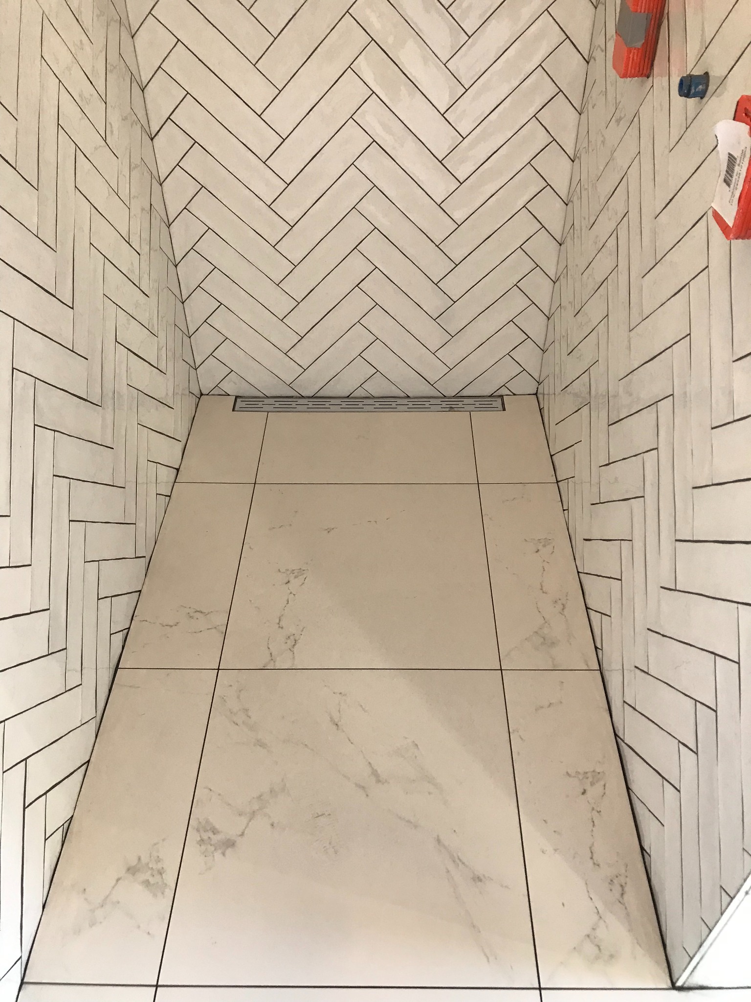 Wykończony brodzik z jasnymi płytkami imitującymi marmur na podłodze i białymi, ułożonymi w jodełkę na ścianach. Widoczny odpływ liniowy.