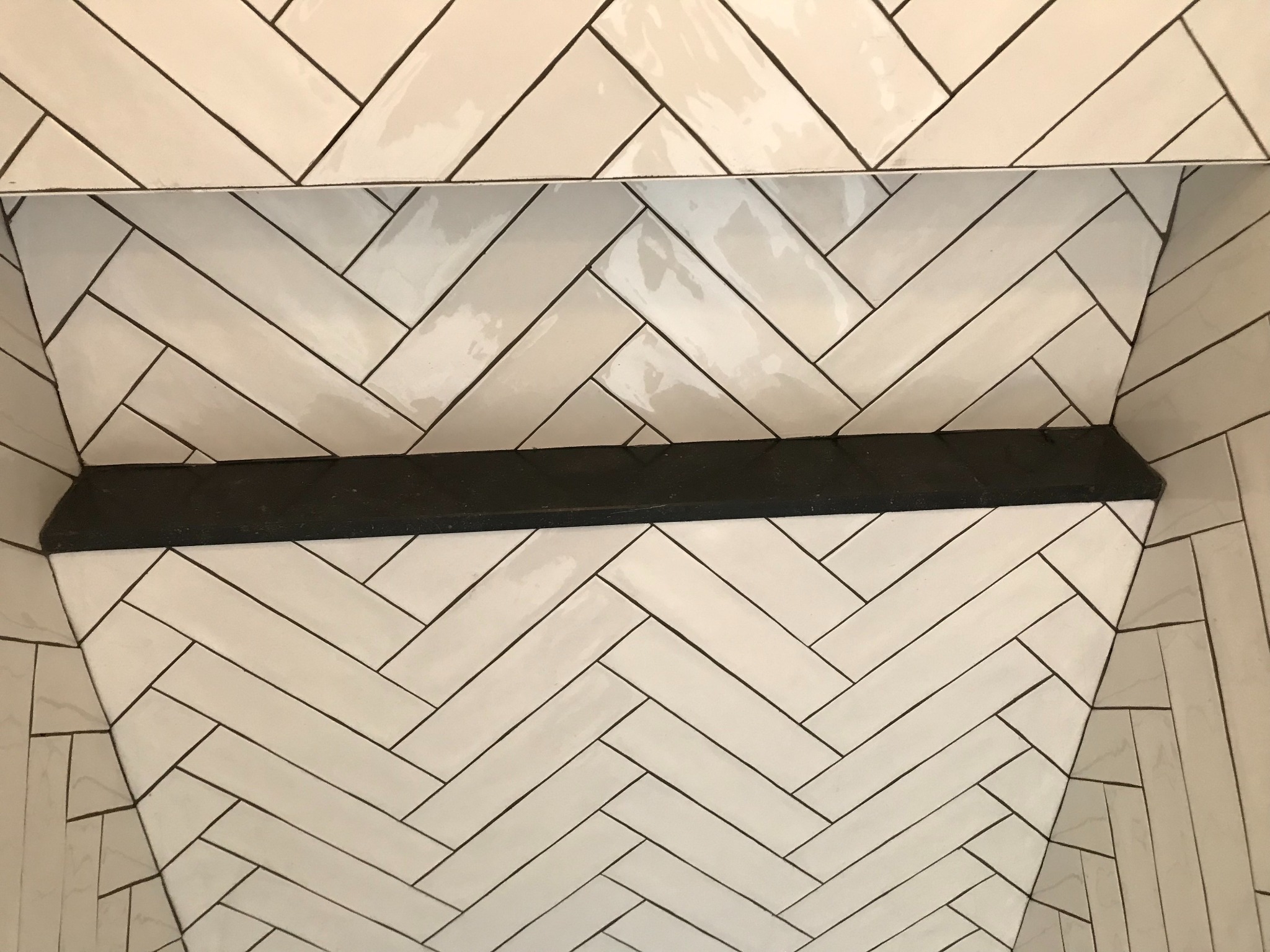 Nisza w łazience wyłożona białymi, podłużnymi płytkami ułożonymi w jodełkę, z czarną półką. Widok z dołu, podkreślający wzór i precyzję wykonania.