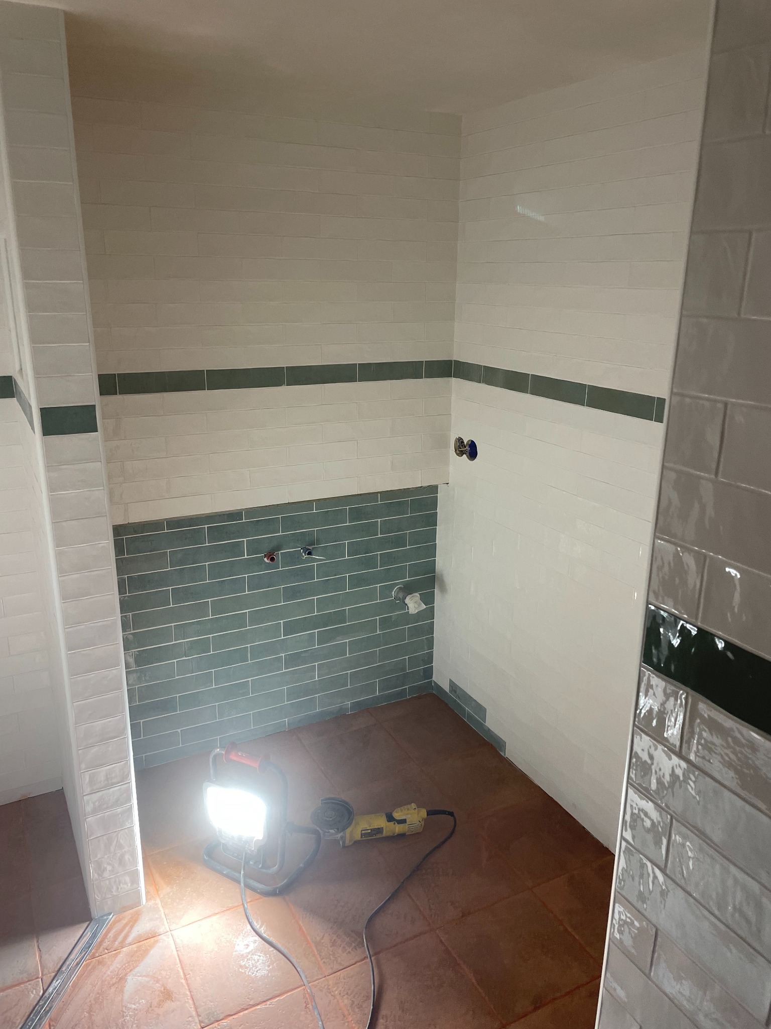 Wnętrze łazienki w trakcie prac wykończeniowych. Widoczne płytki w odcieniach bieli i zieleni, narzędzia oraz lampa budowlana. Ujęcie z niskiej perspektywy.