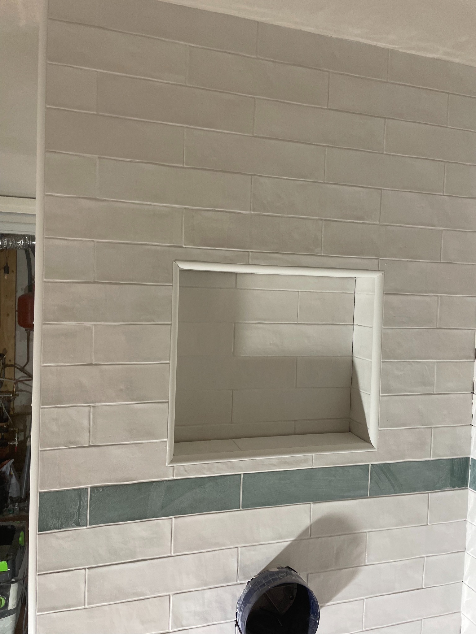 Ściana wyłożona podłużnymi, jasnymi płytkami z wnęką. U dołu widoczny pas ciemniejszych, zielonych płytek. Częściowo widoczny odkurzacz przemysłowy.