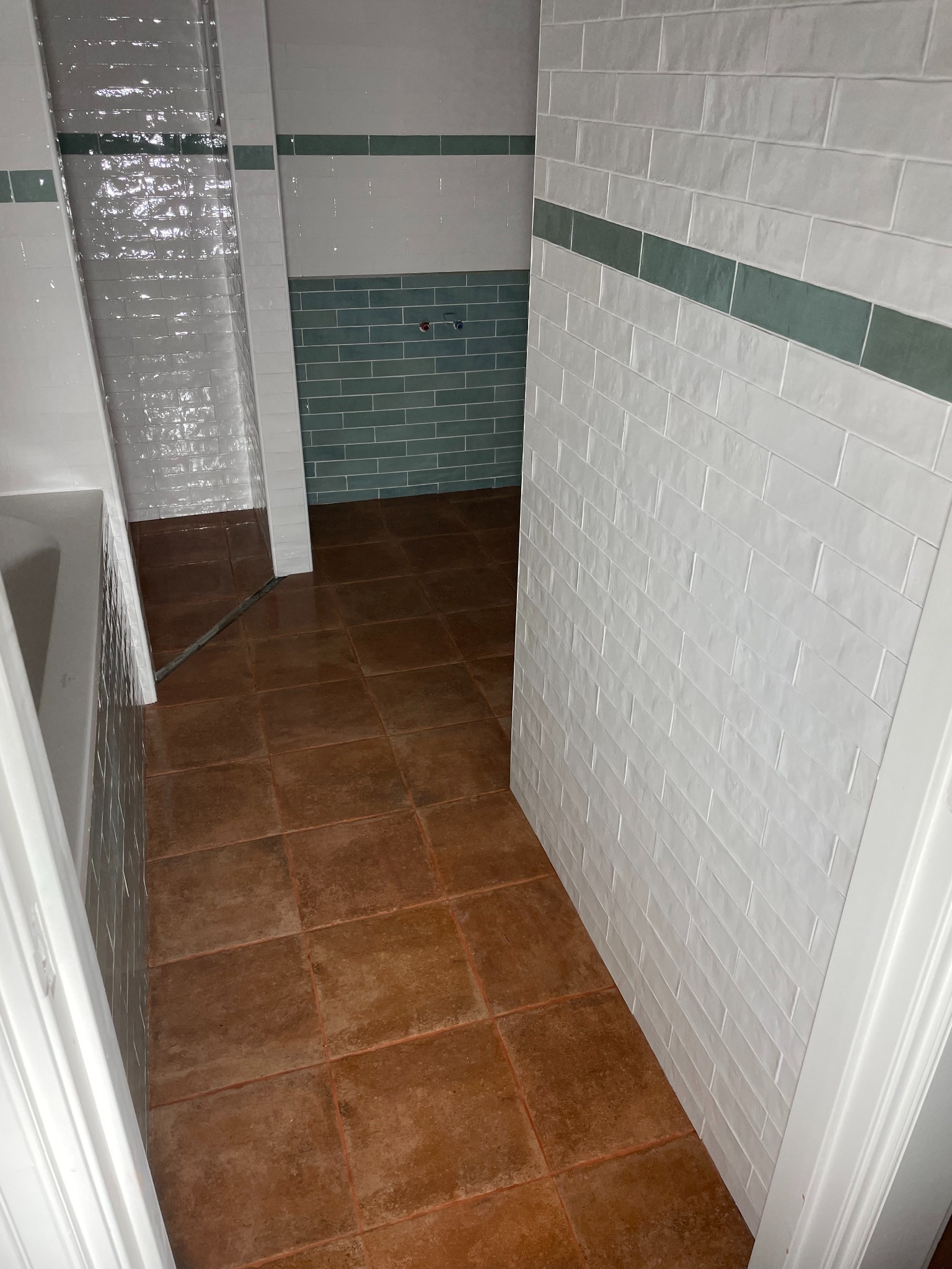 Wykończona łazienka z brązową terakotą i białymi płytkami z zielonym pasem. Widok zza progu, ukazujący kabinę prysznicową i fragment ściany z instalacją.