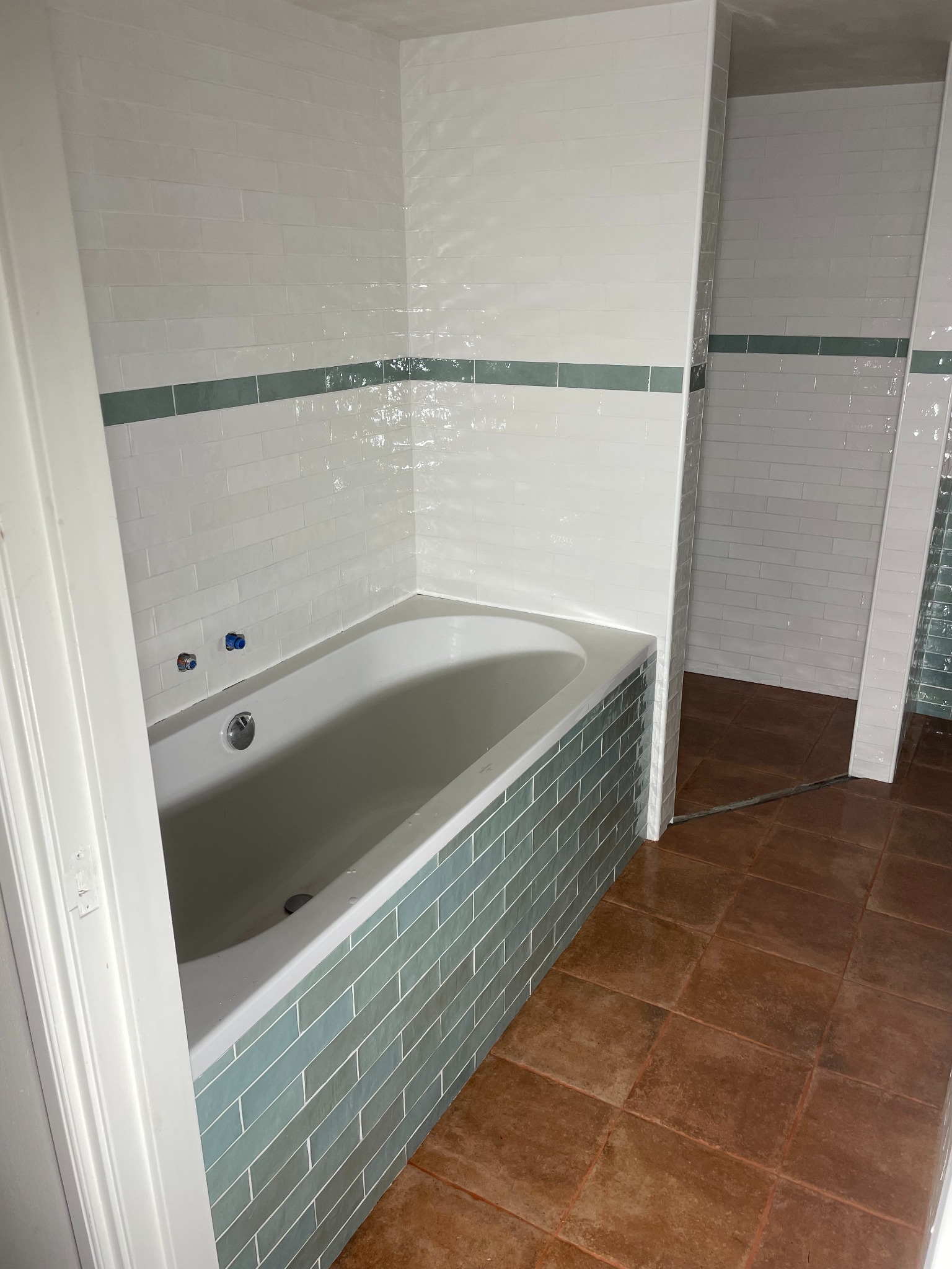 Wykończona łazienka z wanną obłożoną turkusową glazurą, białe płytki na ścianach z zielonym pasem, brązowa podłoga. Widok na dalszą część łazienki.