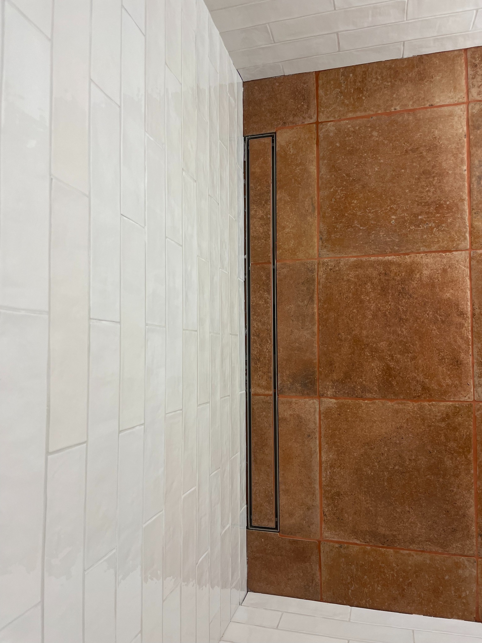 Białe, błyszczące, pionowo ułożone płytki ścienne kontrastują z brązową, kwadratową podłogą. Dyskretny odpływ liniowy wkomponowany w podłogę. Ukończona realizacja.