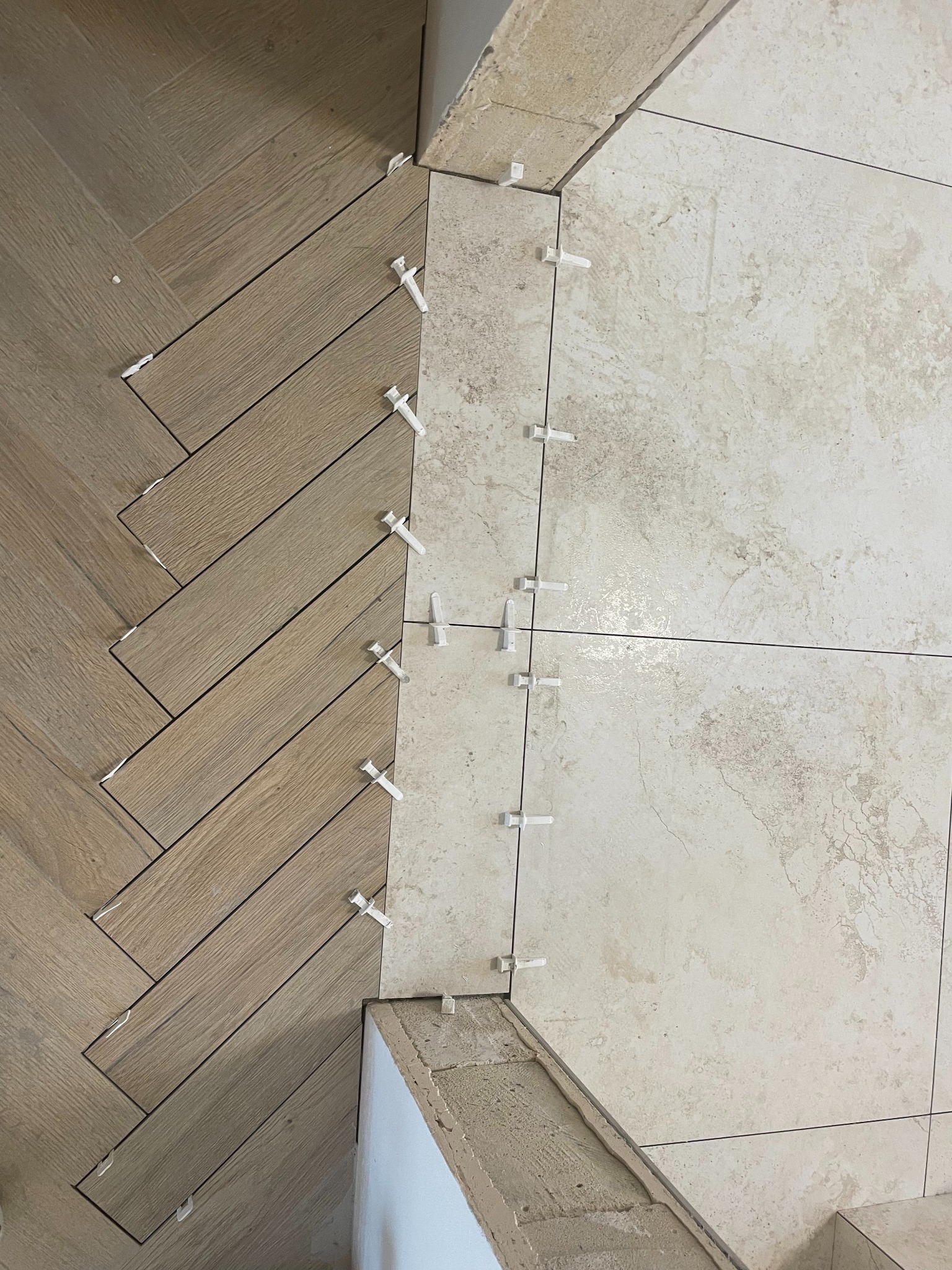 Układanie płytek podłogowych: wzór jodełki z drewnopodobnych płytek łączy się z kwadratowymi płytkami w odcieniach beżu, z użyciem białych krzyżyków dystansowych.