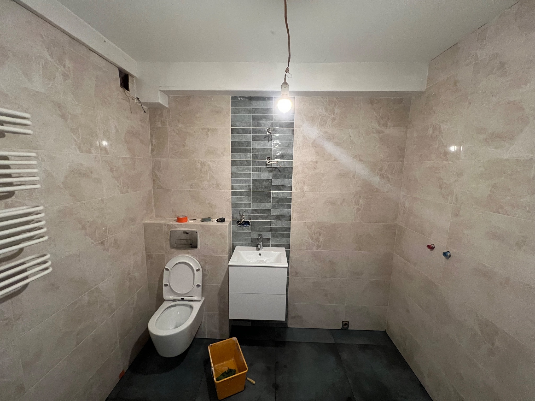 Wykończona łazienka z beżowymi i szarymi płytkami, wiszącą toaletą i umywalką. Widoczny grzejnik drabinkowy i żółte wiadro. Żarówka zwisa z sufitu.