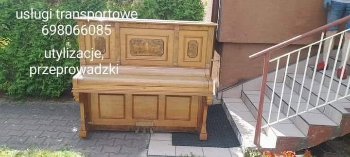 Antyczny, drewniany fortepian przed schodami. Widoczne napisy 'usługi transportowe, utylizacje, przeprowadzki' i numer telefonu. Ujęcie z dołu, zieleń w tle.