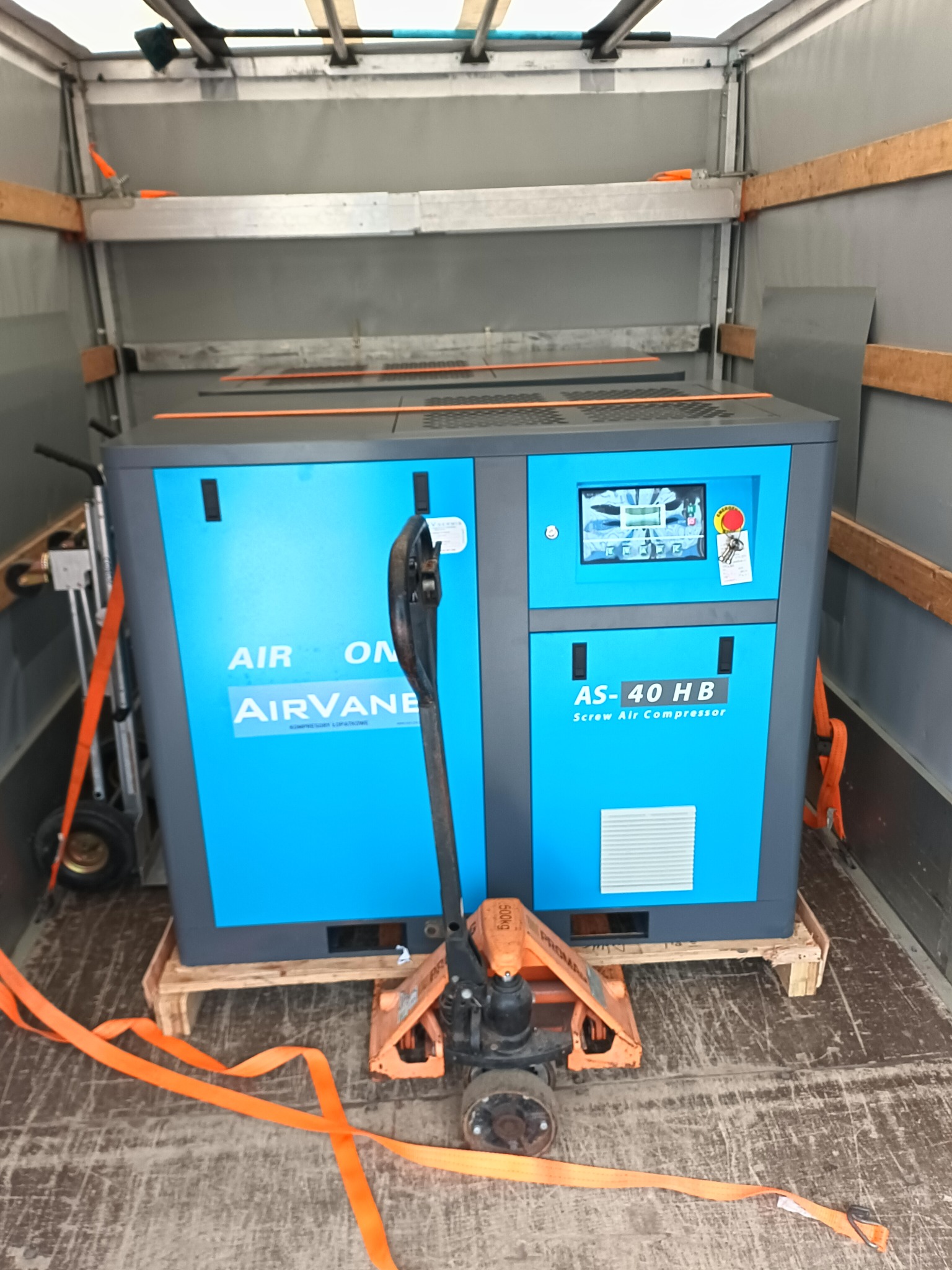Sprężarka śrubowa Airvane AS-40 HB na palecie, zabezpieczona pasami transportowymi wewnątrz przestrzeni ładunkowej busa. Widoczny wózek paletowy.