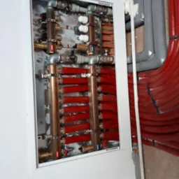 Rozdzielacz ogrzewania podłogowego z rurami w kolorze czerwonym, umieszczony w szafce w ścianie, widoczne liczne zawory i złączki.