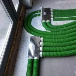 Zielone, karbowane rury instalacyjne prowadzone wzdłuż okna, podłączone do metalowej puszki rozdzielczej na surowym, szarym podłożu. Widoczne pianka montażowa wokół okna.