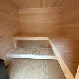 Wood brother - Wnętrze sauny z jasnego drewna: ściany, sufit i dwupoziomowe ławki. Widoczne oświetlenie i piec. Naturalne wykończenie, proste linie, surowy styl.