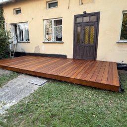 Wood brother - Nowy, drewniany taras przylegający do domu z brązowymi drzwiami. Widoczne deski tarasowe, trawnik i fragment elewacji. Ujęcie z poziomu gruntu.