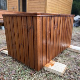 Wood brother - Kwadratowa, drewniana donica w odcieniach brązu, z widocznymi śladami świeżego malowania, ustawiona na trawie przed drewnianym budynkiem. Detal wykończenia.