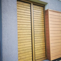 Wood brother - Drewniana zabudowa okienna z poziomych desek, przylegająca do ściany budynku o szarej, chropowatej fakturze. Obok widoczna zabudowa z paneli w odcieniach brązu.