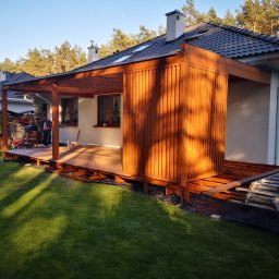 Wood brother - Drewniana konstrukcja tarasu z elementami zabudowy pionowej, widoczna w trakcie montażu. Na pierwszym planie trawnik, w tle dom jednorodzinny i drzewa.