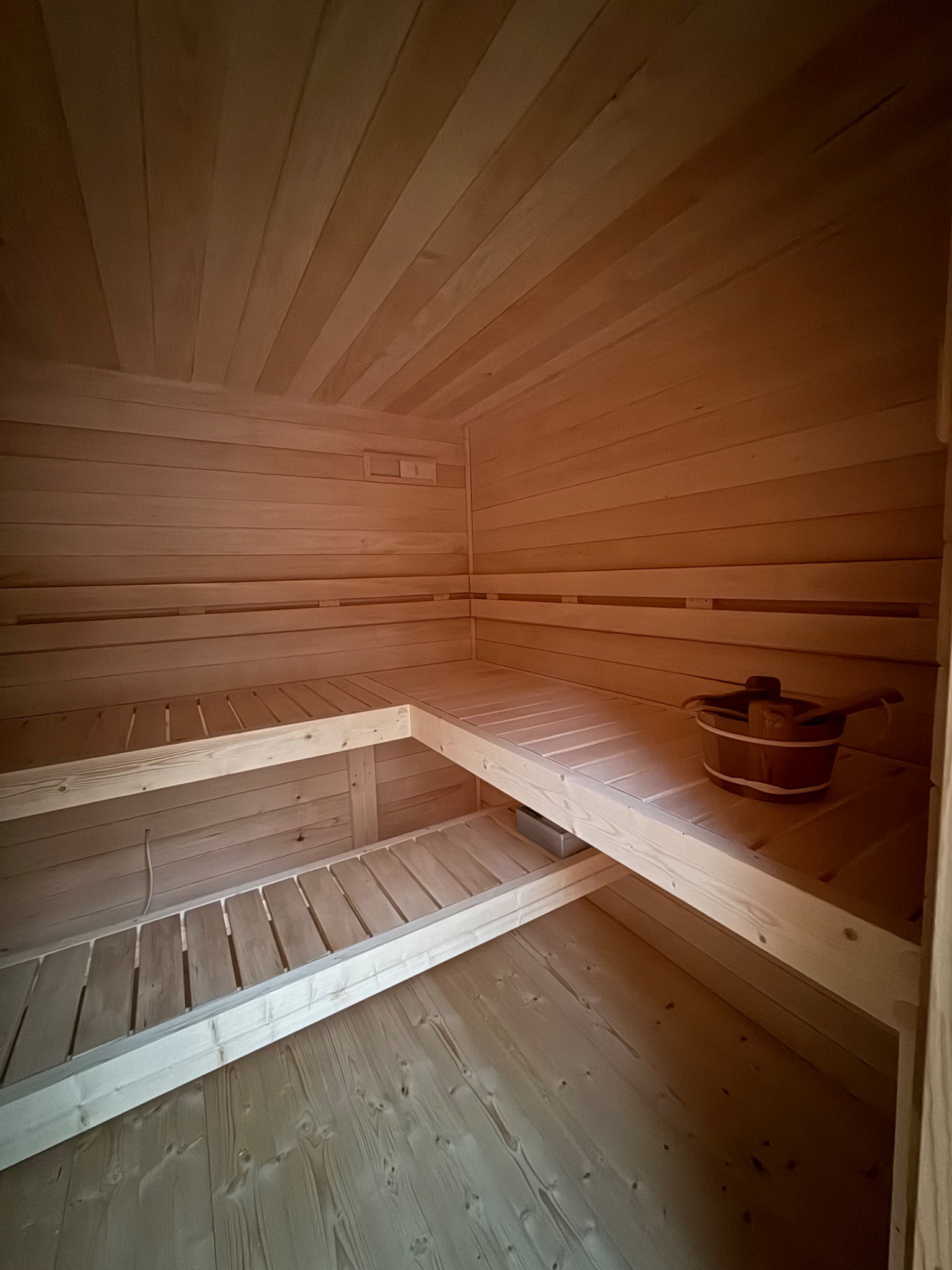 Jasne drewno w saunie: ławy na dwóch poziomach, wiadro z chochlą. Ściany i sufit wyłożone boazerią. Minimalistyczny design, ciepłe oświetlenie.