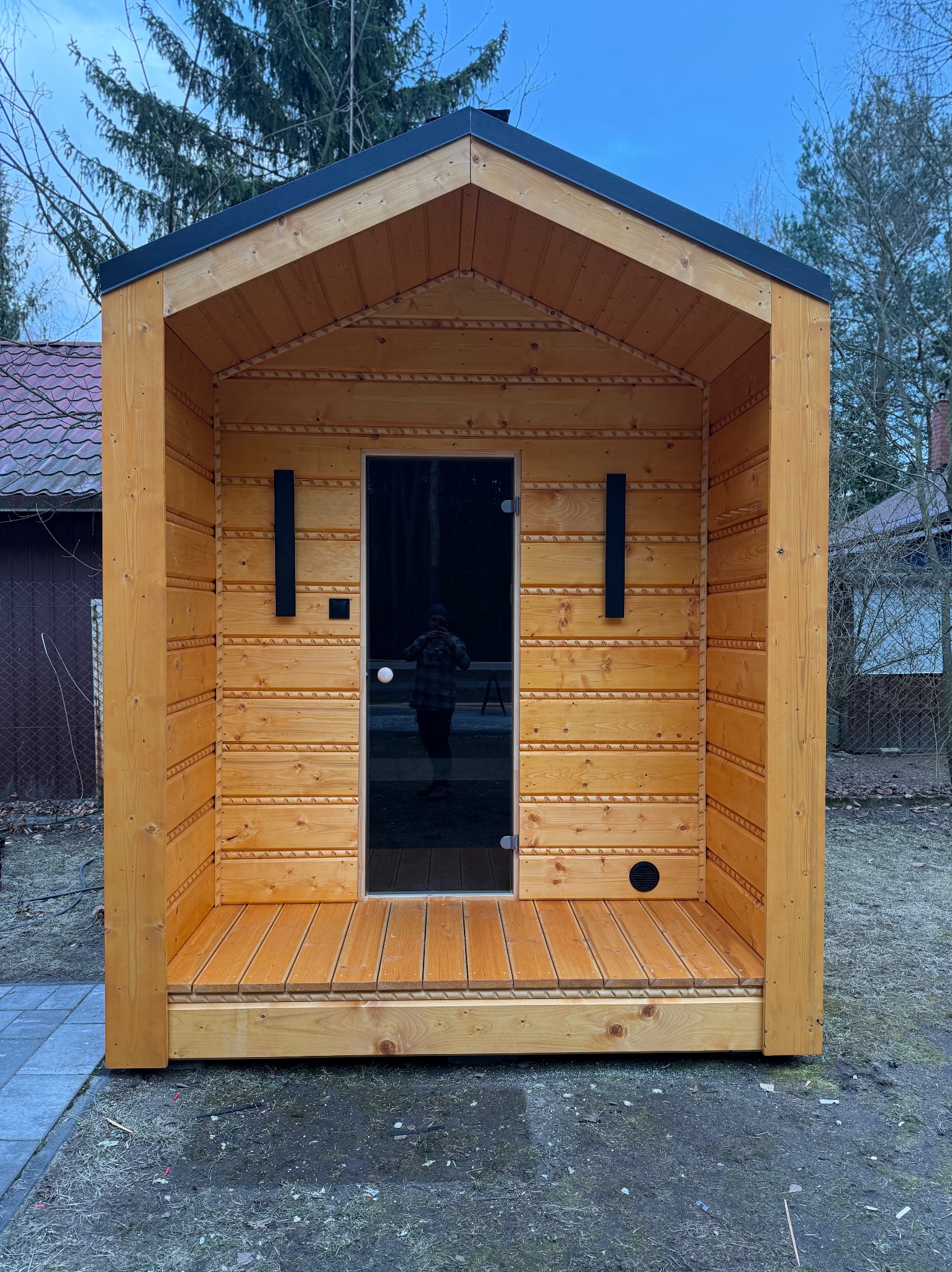 Drewniana, wolnostojąca konstrukcja z przeszklonymi drzwiami, przypominająca małą saunę lub domek ogrodowy. Widoczne detale wykończenia i oświetlenie zewnętrzne.