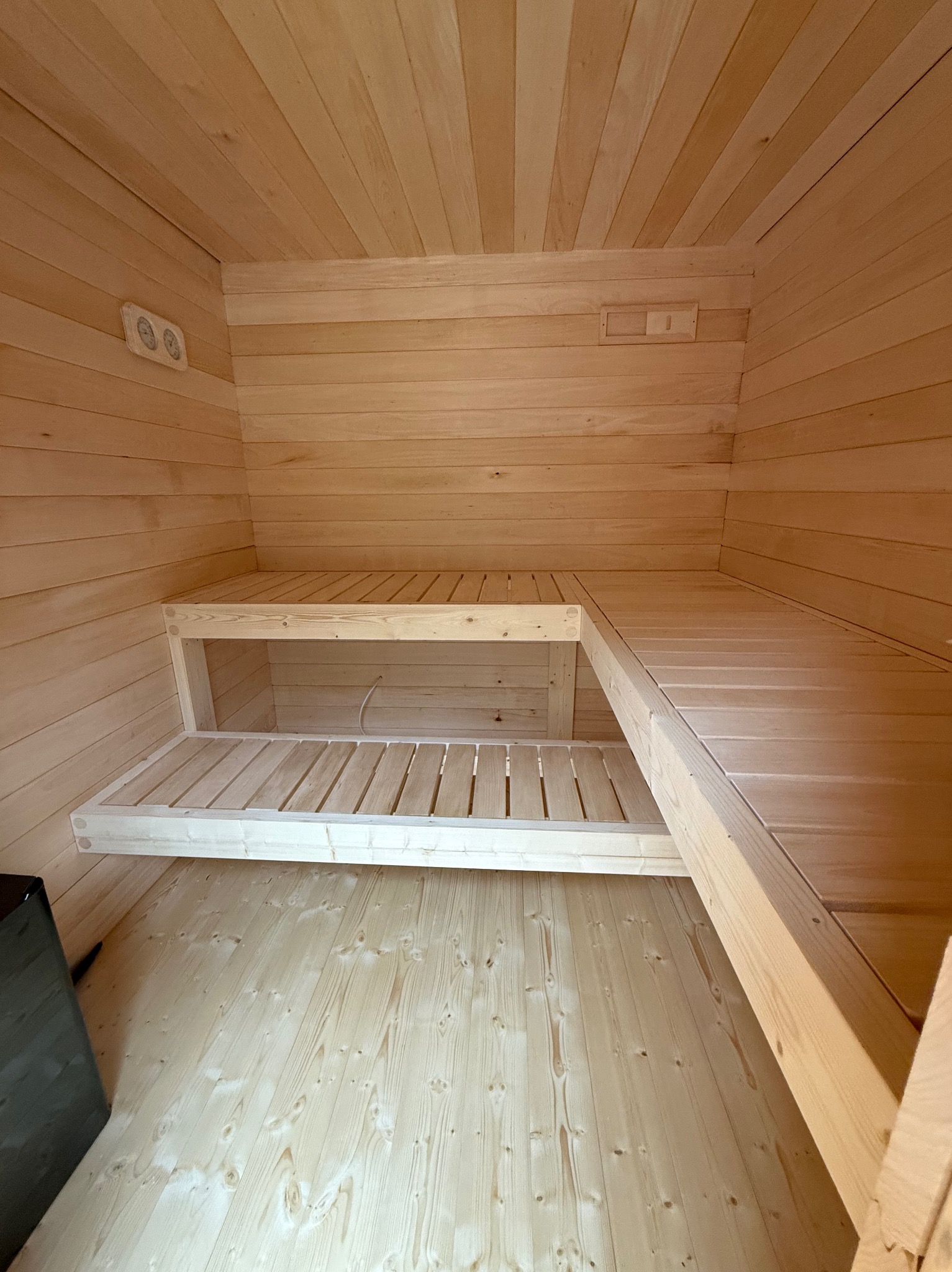 Wnętrze sauny z jasnego drewna: ściany, sufit i dwupoziomowe ławki. Widoczne oświetlenie i piec. Naturalne wykończenie, proste linie, surowy styl.