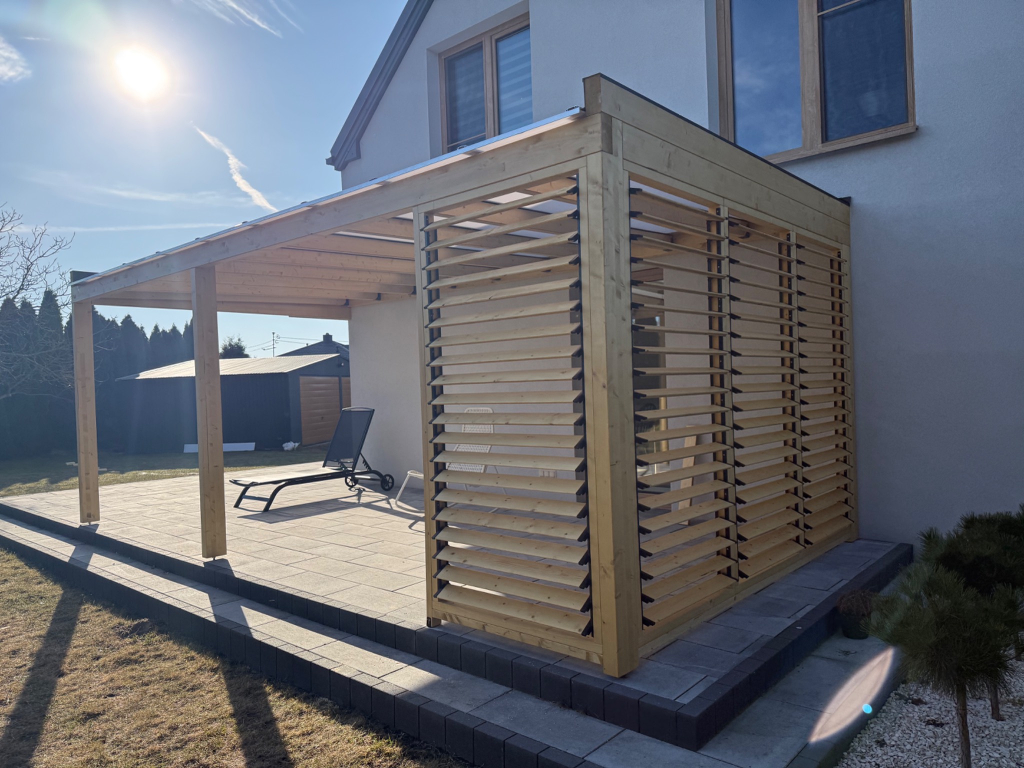 Drewniana pergola z żaluzjowymi ścianami, przylegająca do domu, z widocznym tarasem z szarej kostki. W tle zieleń i błękitne niebo z delikatną smugą kondensacyjną.