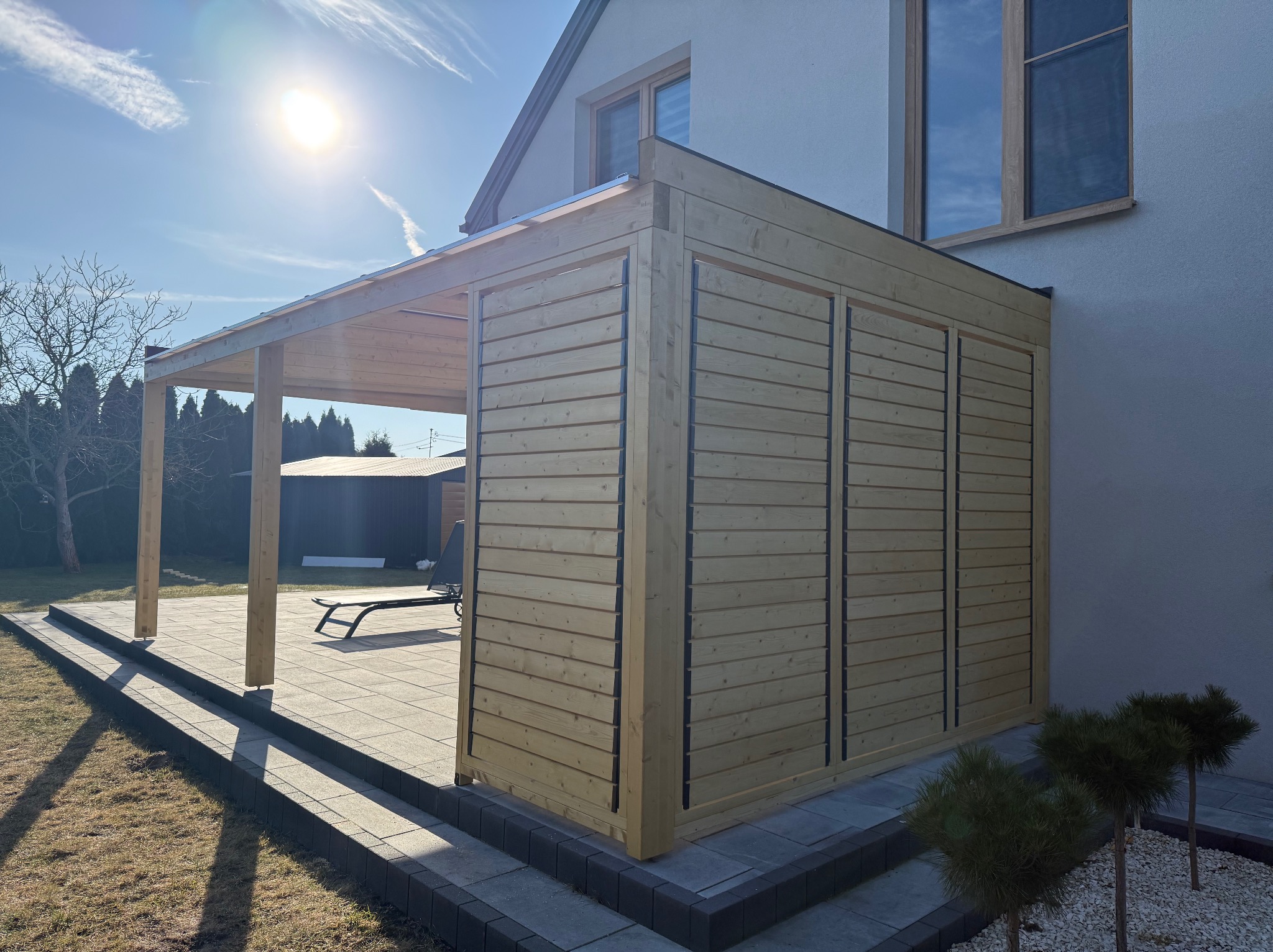 Drewniana pergola z zabudową, przylegająca do domu, z widocznym tarasem z kostki. Jasne drewno, poziome deski, nowoczesny design. W tle leżak.