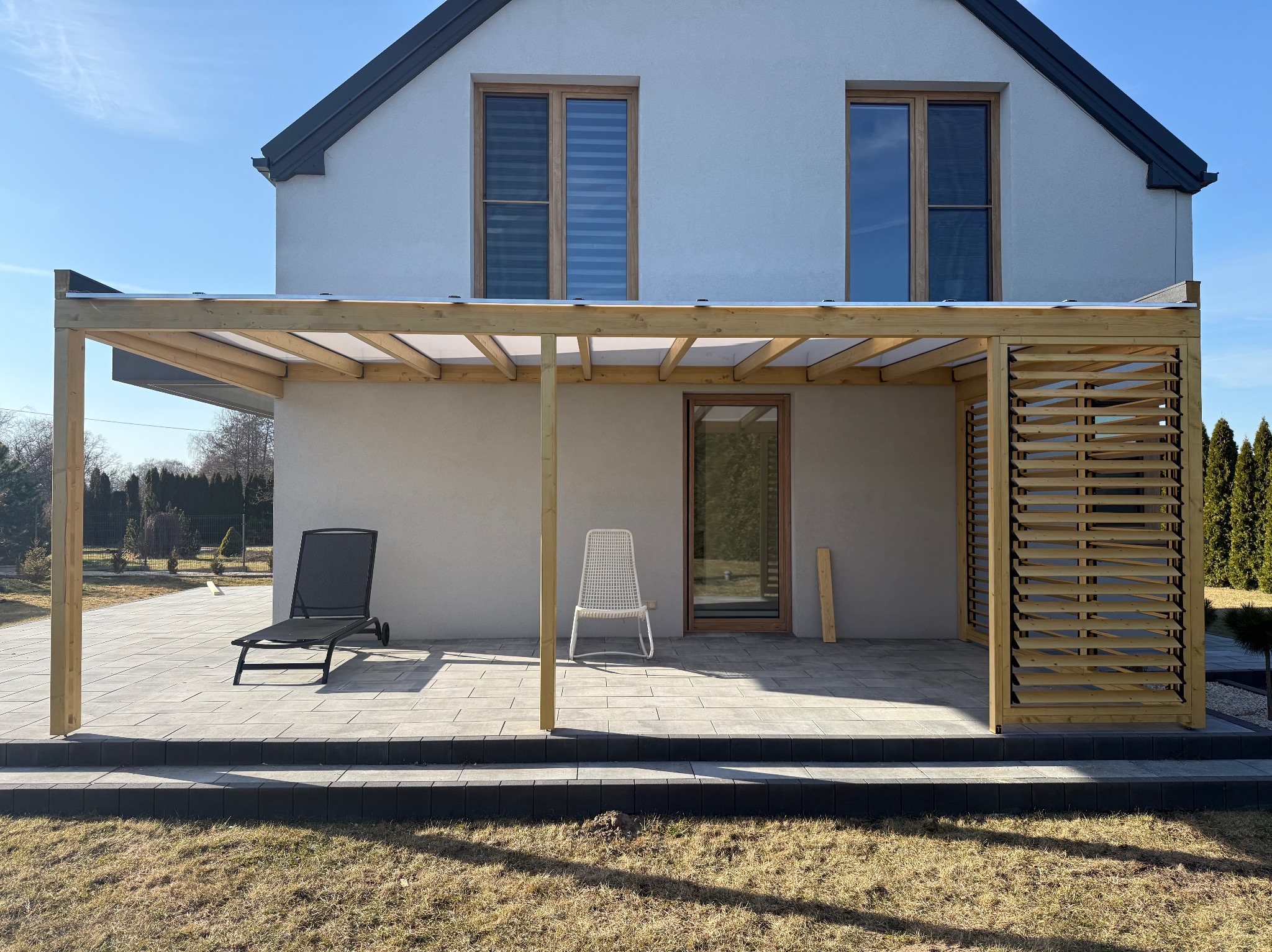 Nowoczesna pergola drewniana z zadaszeniem na tarasie z szarej kostki, z leżakiem i krzesłem. Widok na dom z szarą elewacją i drewnianymi oknami.