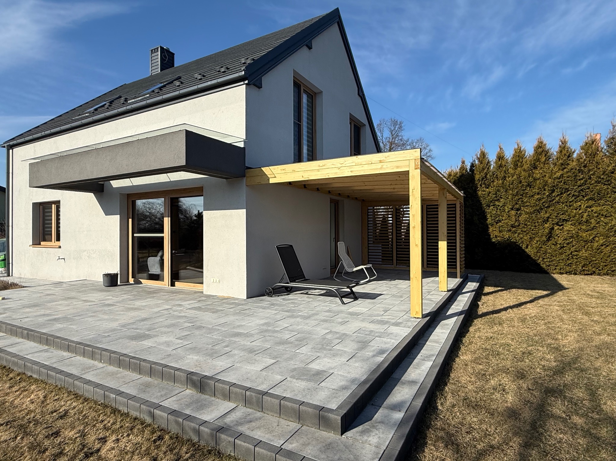 Nowoczesny dom z betonowym tarasem i drewnianą pergolą. Dwa leżaki na tarasie. Tuje w tle. Słoneczny dzień.