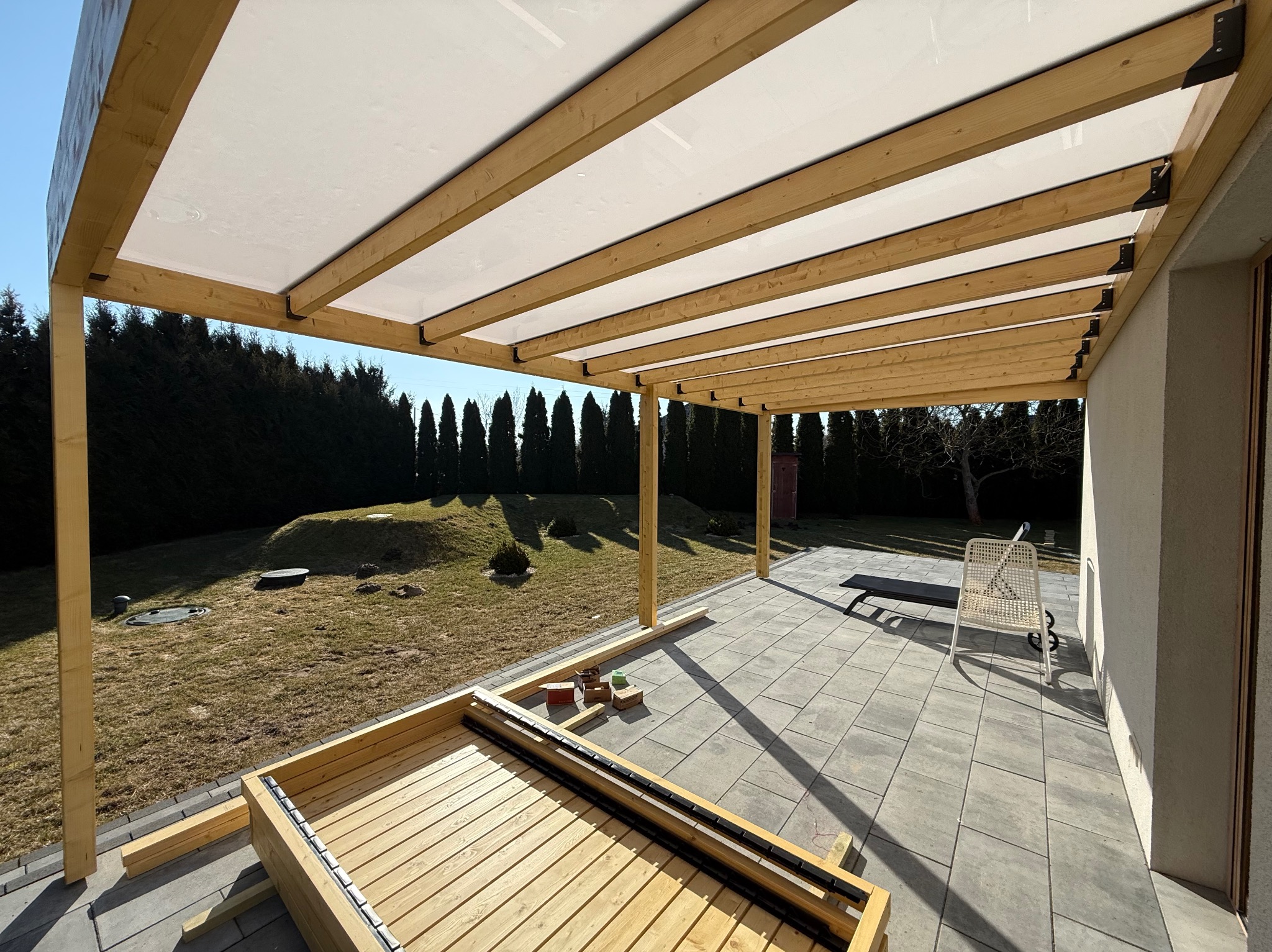 Drewniana pergola z białym zadaszeniem w trakcie montażu, widok na ogród z tujami. Częściowo zmontowany taras z desek na szarych płytach.