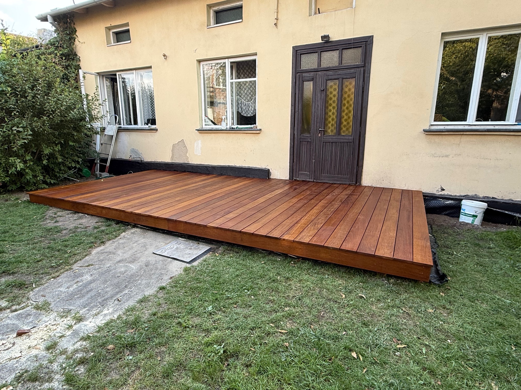 Nowy, drewniany taras przylegający do domu z brązowymi drzwiami. Widoczne deski tarasowe, trawnik i fragment elewacji. Ujęcie z poziomu gruntu.