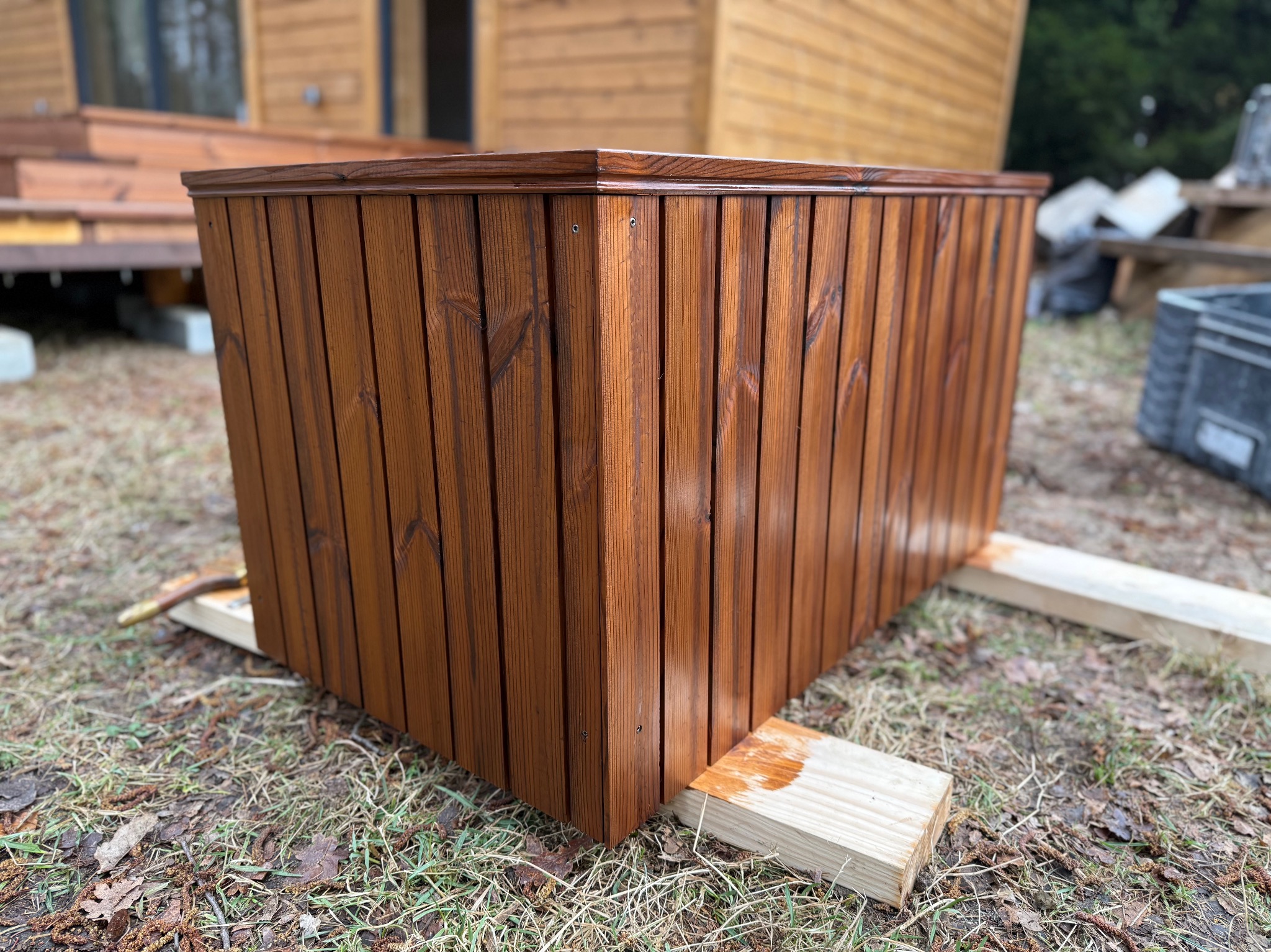 Kwadratowa, drewniana donica w odcieniach brązu, z widocznymi śladami świeżego malowania, ustawiona na trawie przed drewnianym budynkiem. Detal wykończenia.