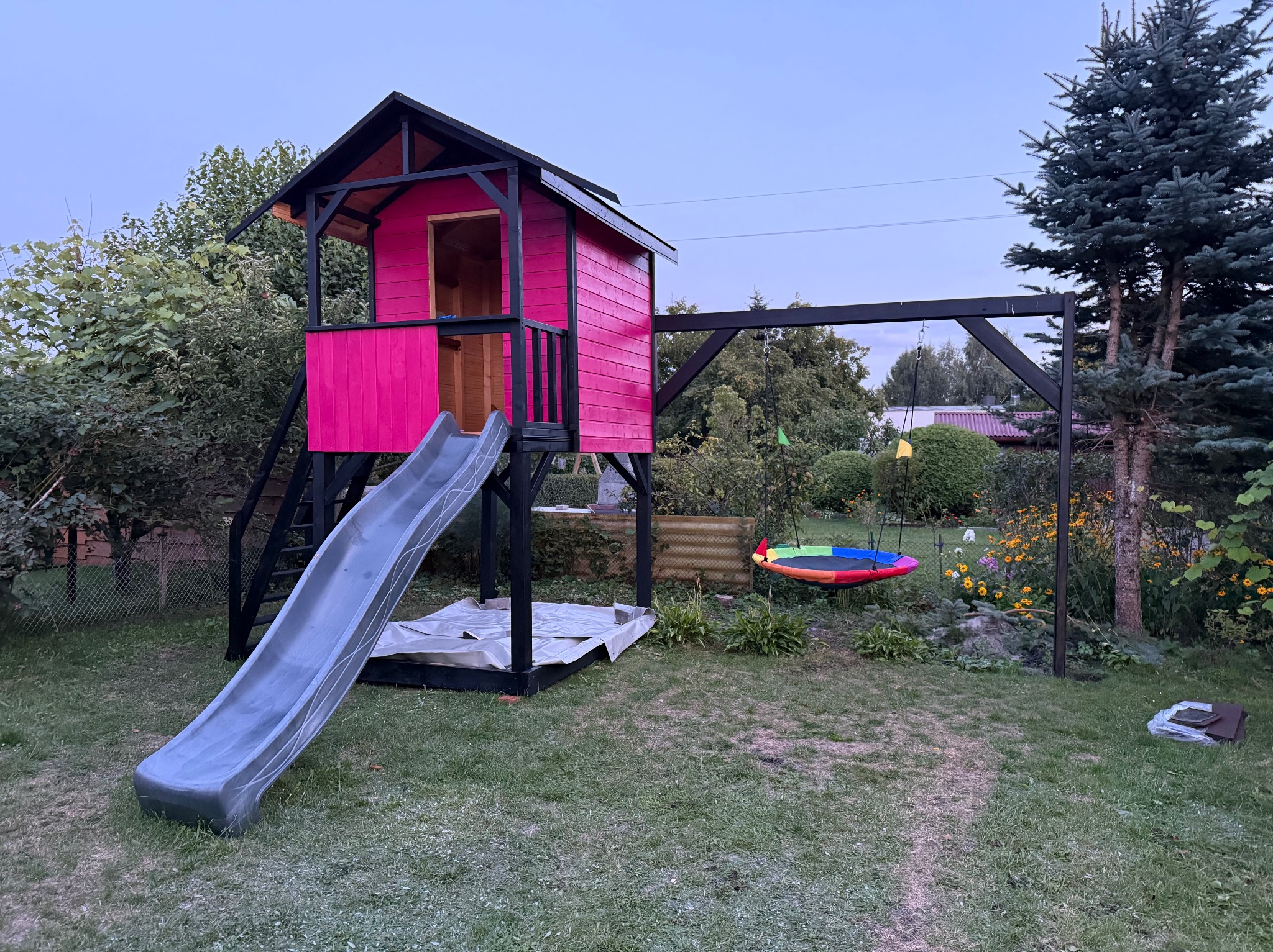 Kolorowy plac zabaw w ogrodzie: różowy domek na palach ze zjeżdżalnią i huśtawka typu bocianie gniazdo na czarnej konstrukcji. Otoczenie to trawa i zieleń.