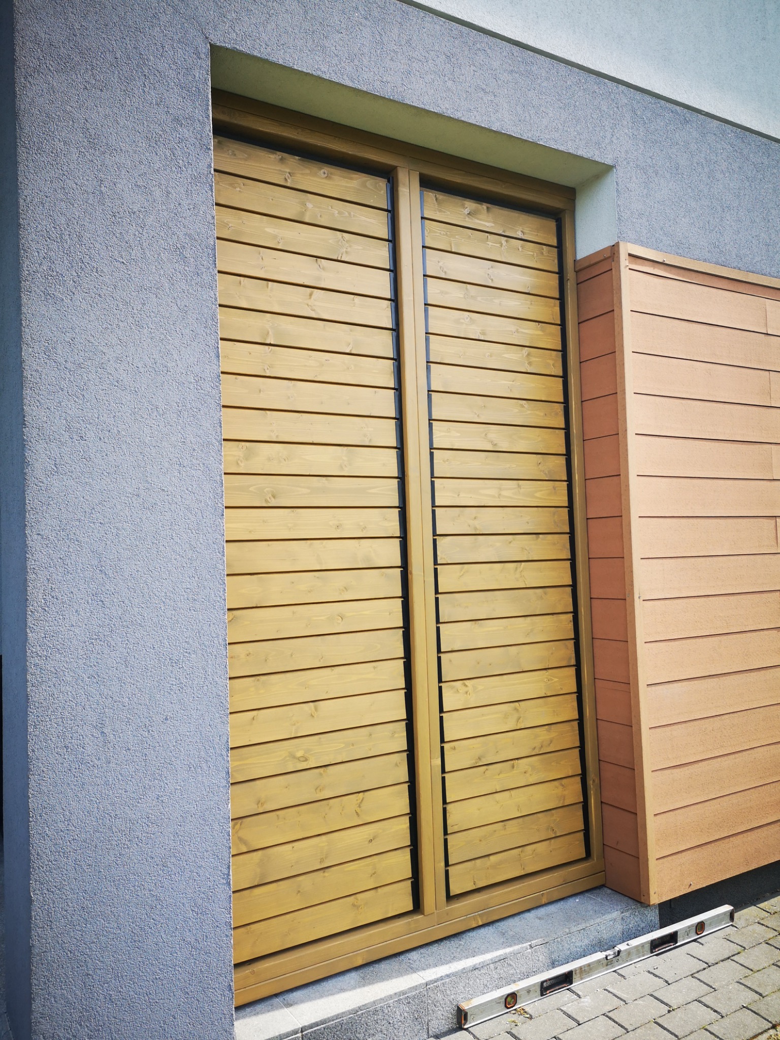 Drewniana zabudowa okienna z poziomych desek, przylegająca do ściany budynku o szarej, chropowatej fakturze. Obok widoczna zabudowa z paneli w odcieniach brązu.