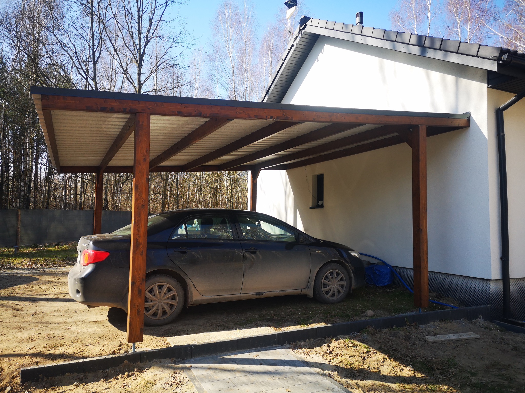 Drewniana wiata garażowa przylegająca do białego domu, z zaparkowanym czarnym samochodem. Konstrukcja z belek, zadaszenie z blachy falistej. Widok w słoneczny dzień.