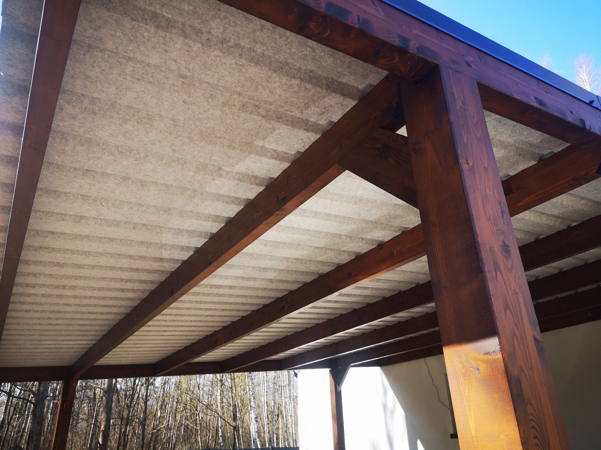 Drewniana konstrukcja z zadaszeniem, widok od dołu. Ciemne, lakierowane belki i jasne zadaszenie tworzą kontrast. W tle drzewa bez liści i fragment błękitnego nieba.